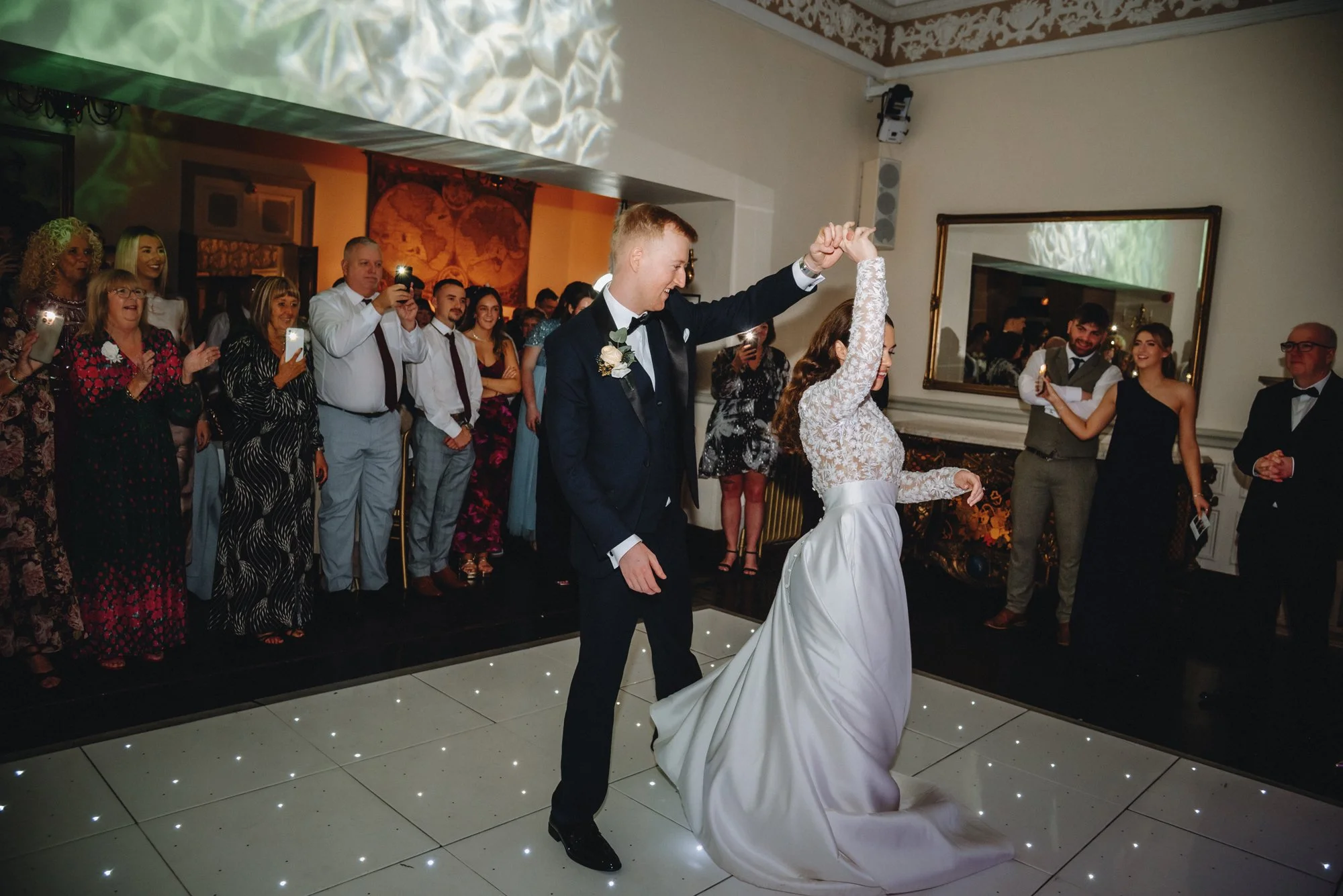 A bride and groom are dancing at their wedding reception, surrounded by guests taking photos and watching.