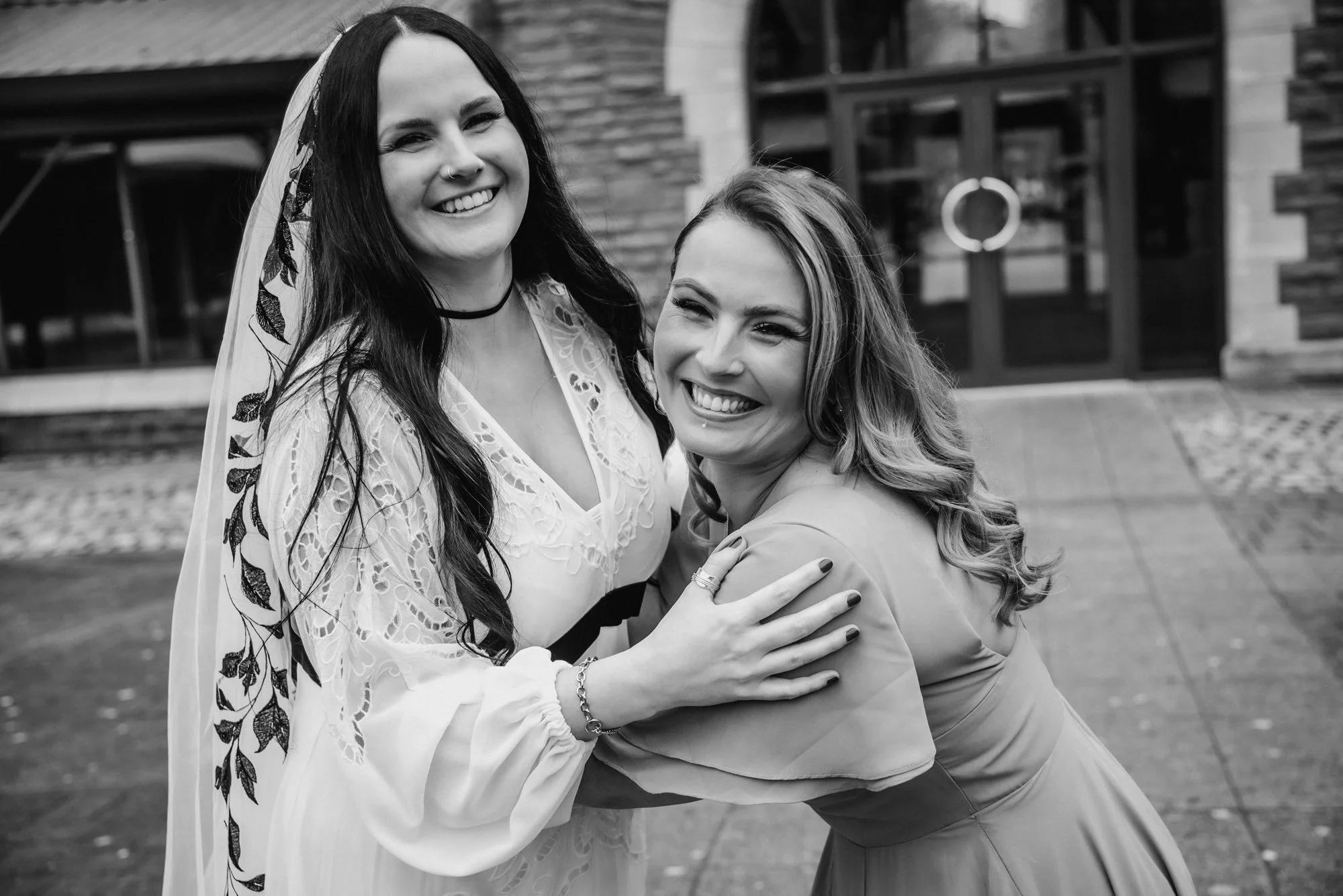 Two women smiling, hugging each other outdoors in front of a building, one wearing a wedding dress and veil, the other in a dress with wavy hair.