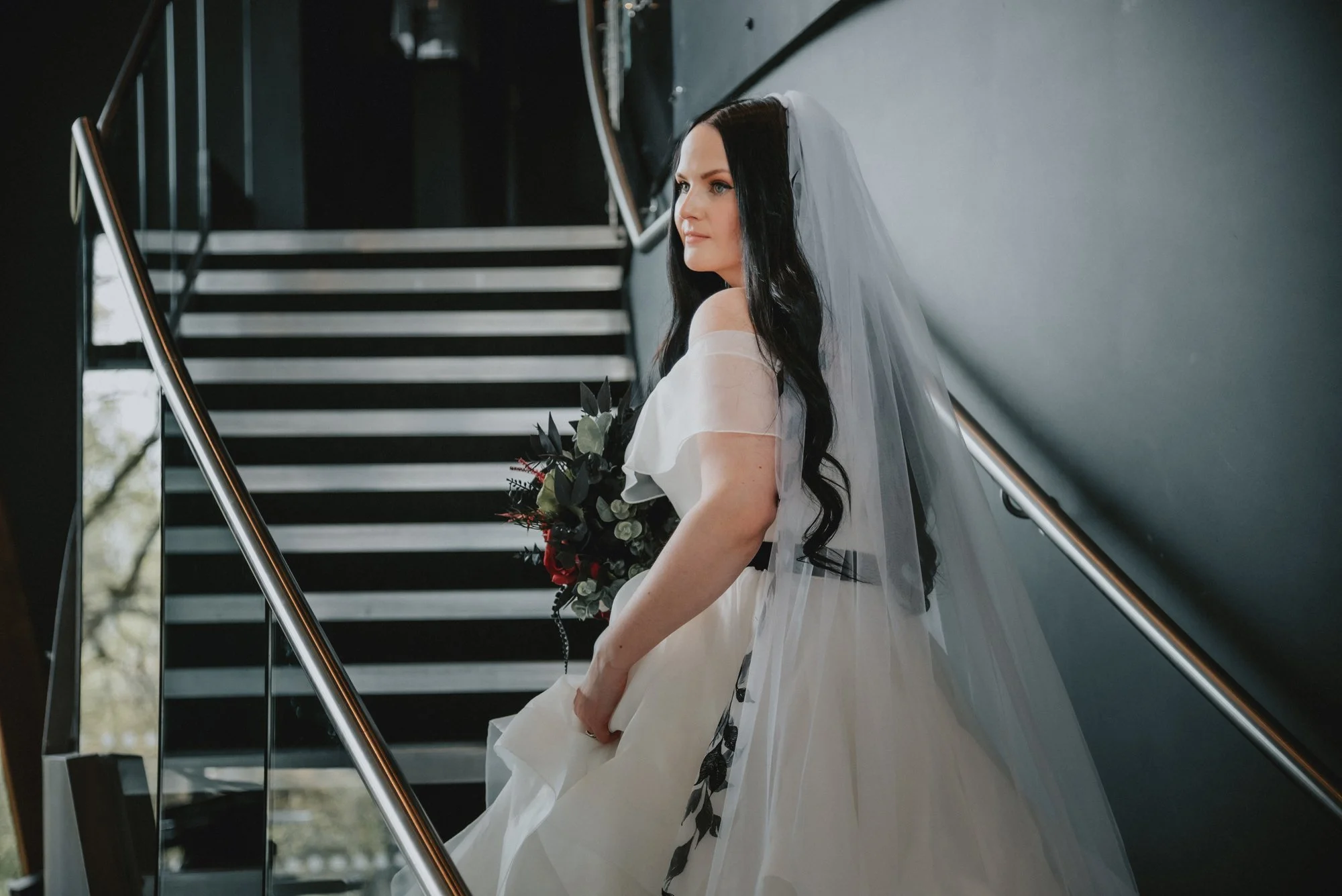 A bride with long dark hair wearing a white off-the-shoulder wedding dress, veil, and holding a bouquet, standing at the bottom of a staircase, looking to the side.