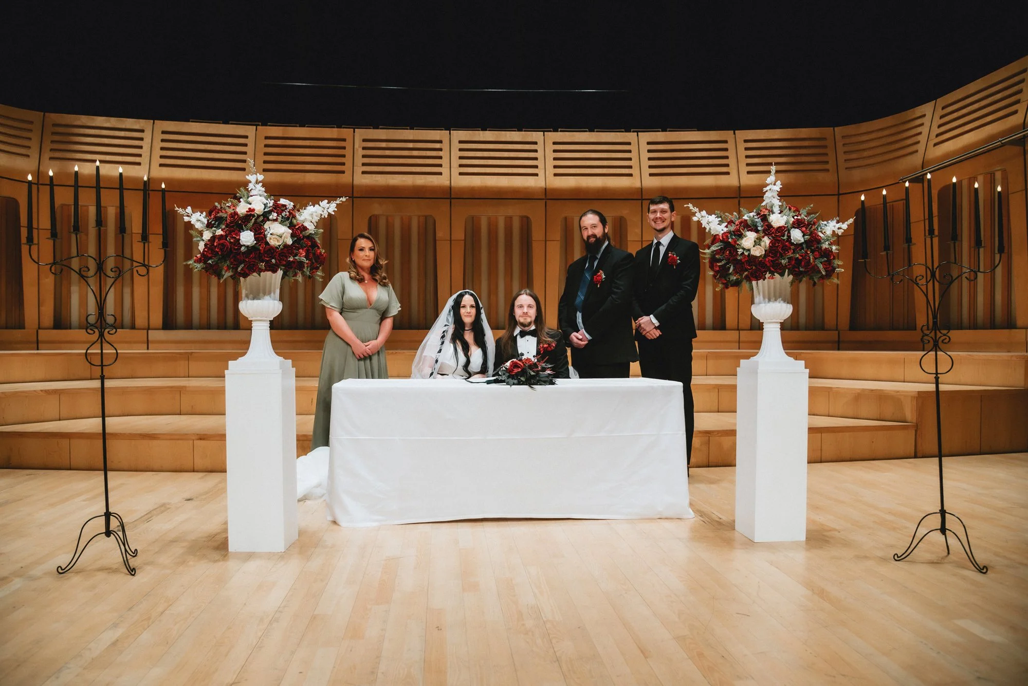 A wedding ceremony in a concert hall with six people, including the bride and groom seated at a table with floral arrangements, surrounded by servers, with large floral displays, candelabras, and a wooden curved wall in the background.