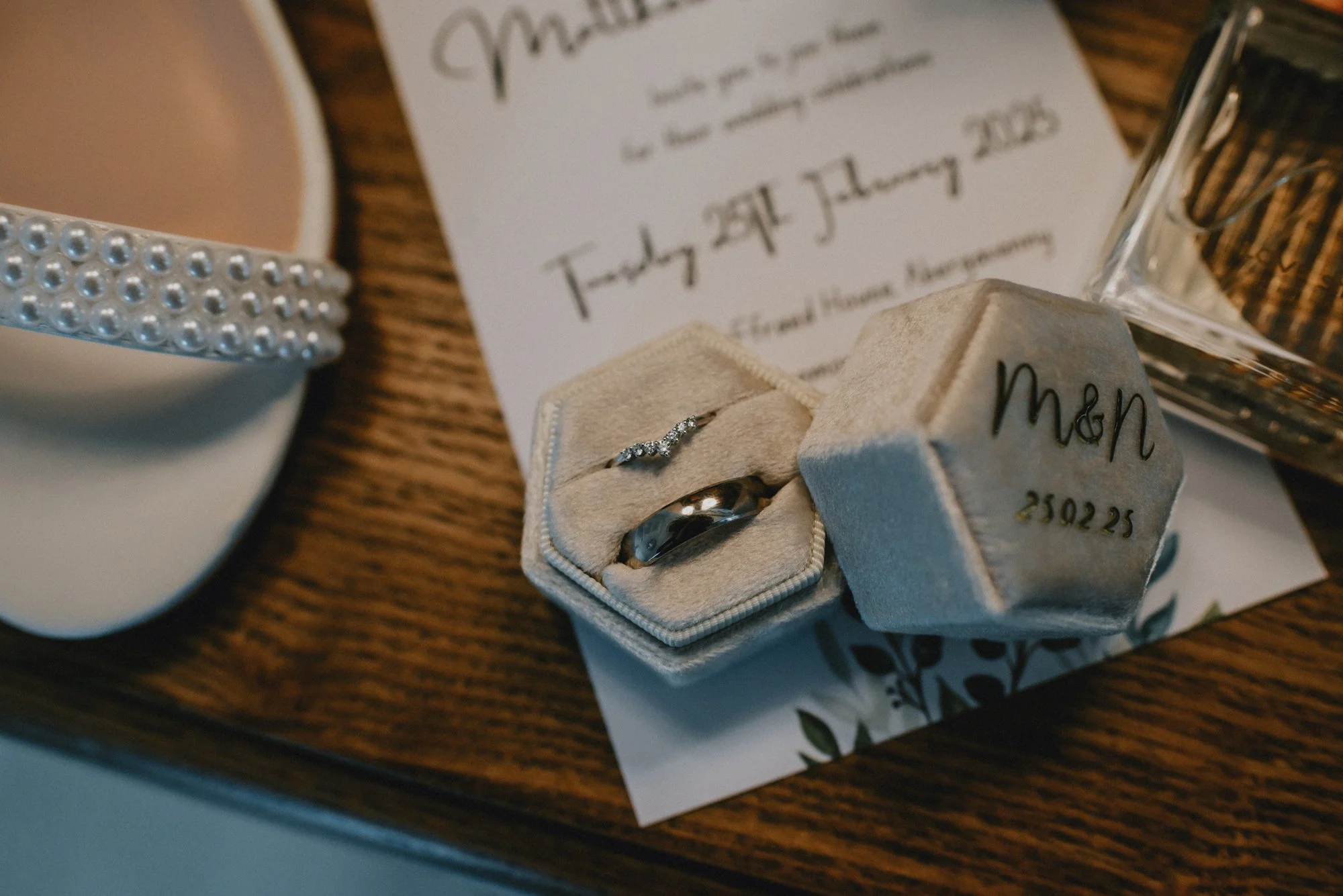 A wedding invitation on a wooden table along with a ring box containing a diamond ring and a wedding band. The ring box has the word 'Man' and a date written on it. There is also a white cup of coffee with a decorative pearl band around it, and a gla