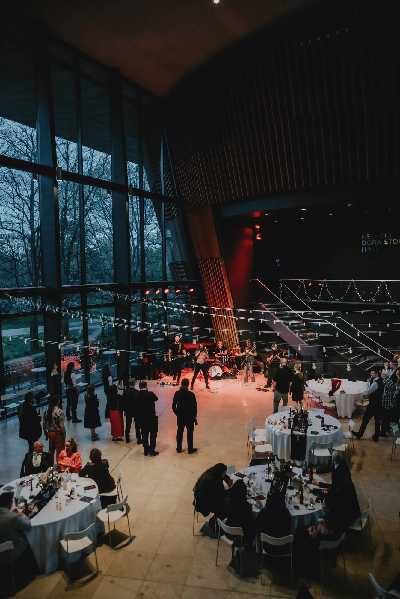 Indoor event at a modern venue with a live band performing on stage. People are standing and dancing near the stage, while others are seated at round tables with white tablecloths. The venue has large floor-to-ceiling windows showing trees outside an