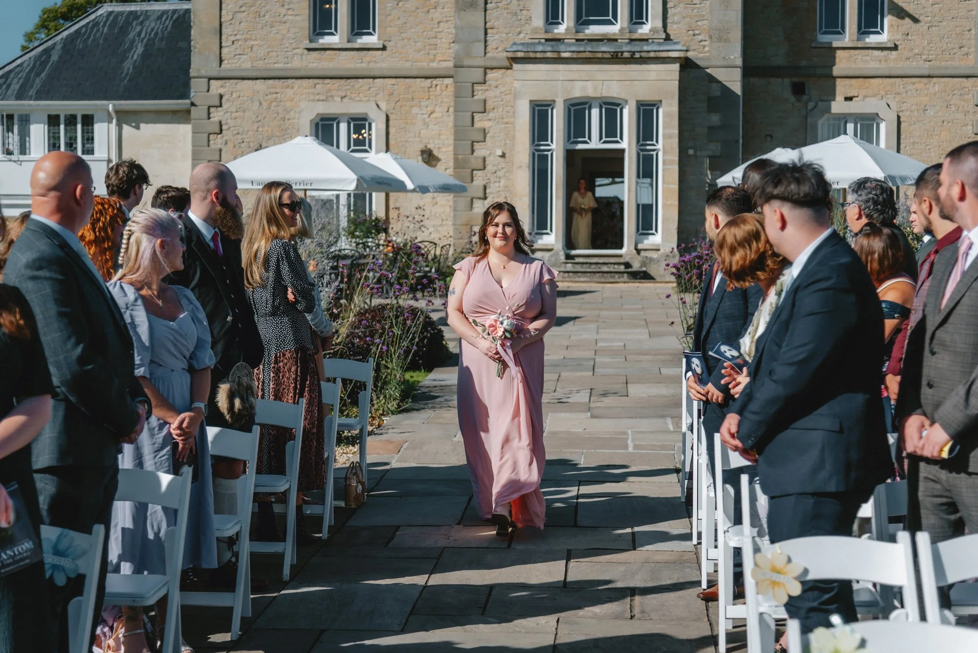A woman in a pink dress walking down the aisle holding a bouquet, at an outdoor wedding ceremony with guests on either side.