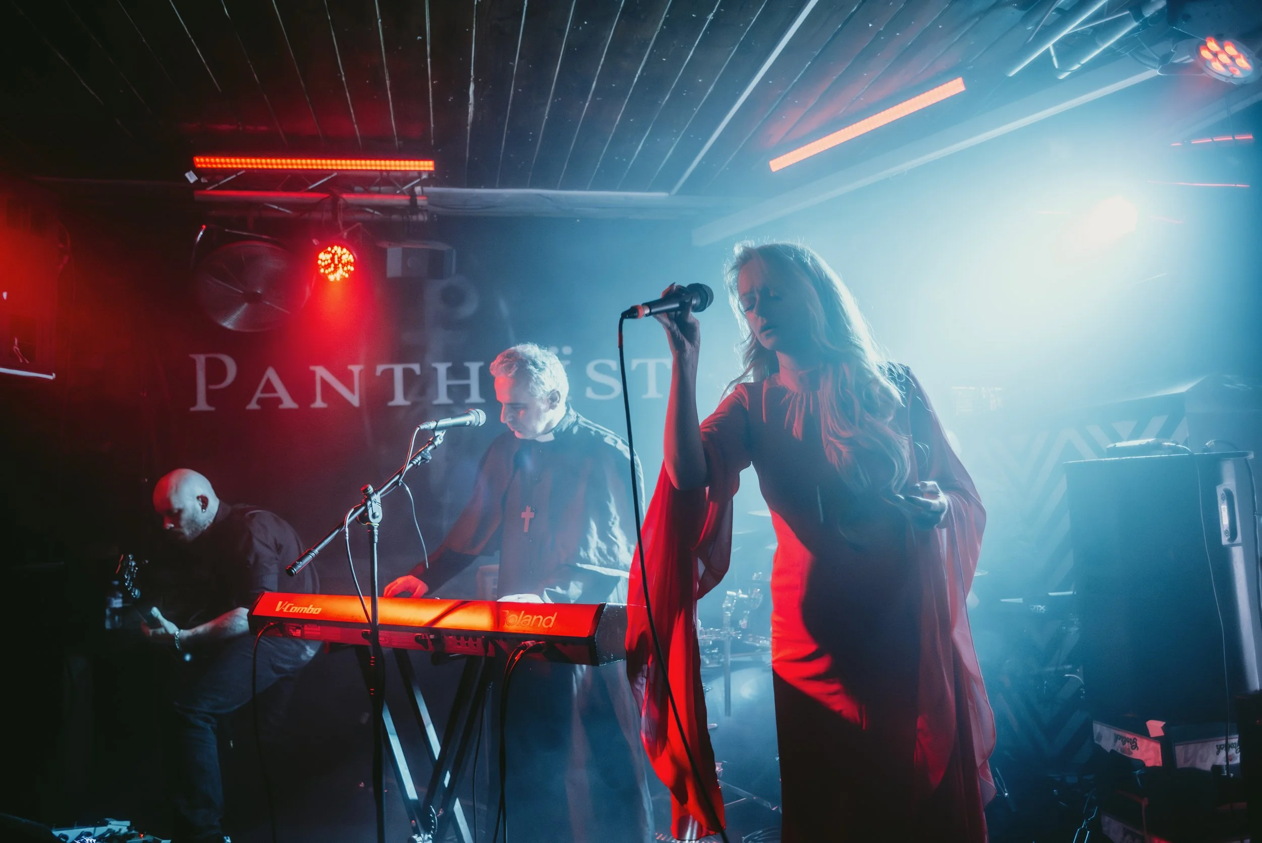 A band performing live on stage with a female singer, a keyboard player, and a guitarist, with stage lights and a sign that reads 'PANTHERS' in the background.