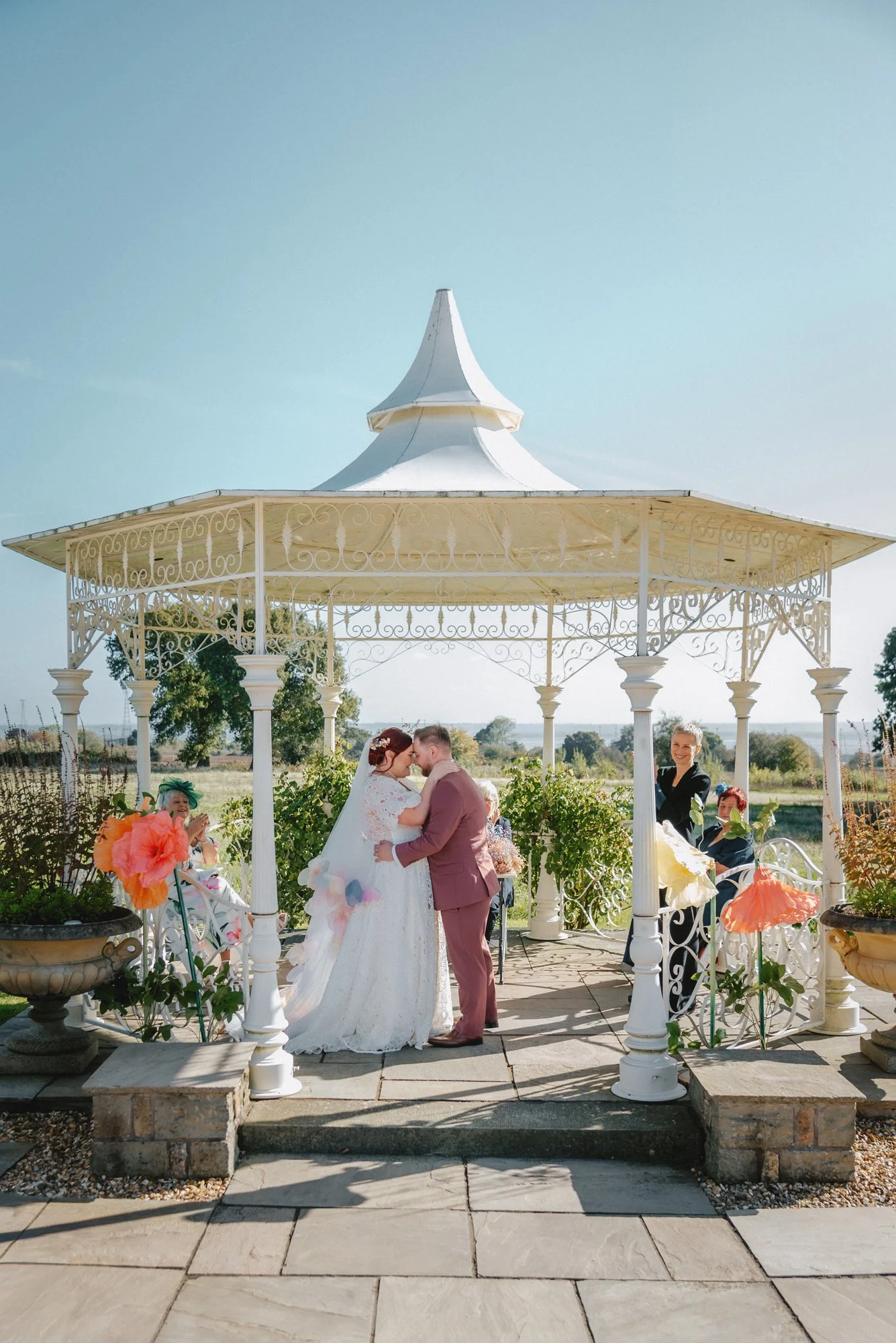 A wedding ceremony taking place in a white gazebo outdoors, with a bride and groom kissing, surrounded by guests seated and clapping, in a garden setting with trees and a clear blue sky.