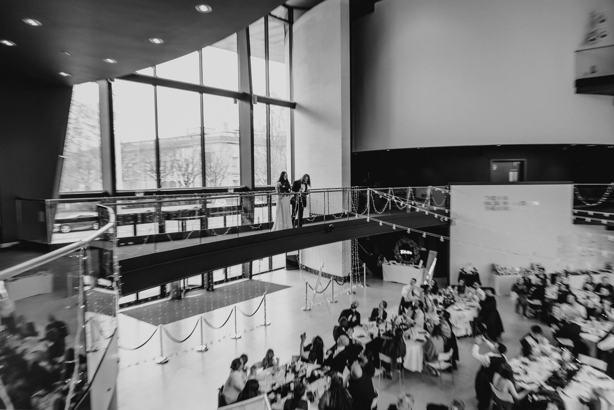 A large indoor event space with many people seated at tables, and two women standing on an upper balcony near a large window, decorated with string lights and festive decorations.