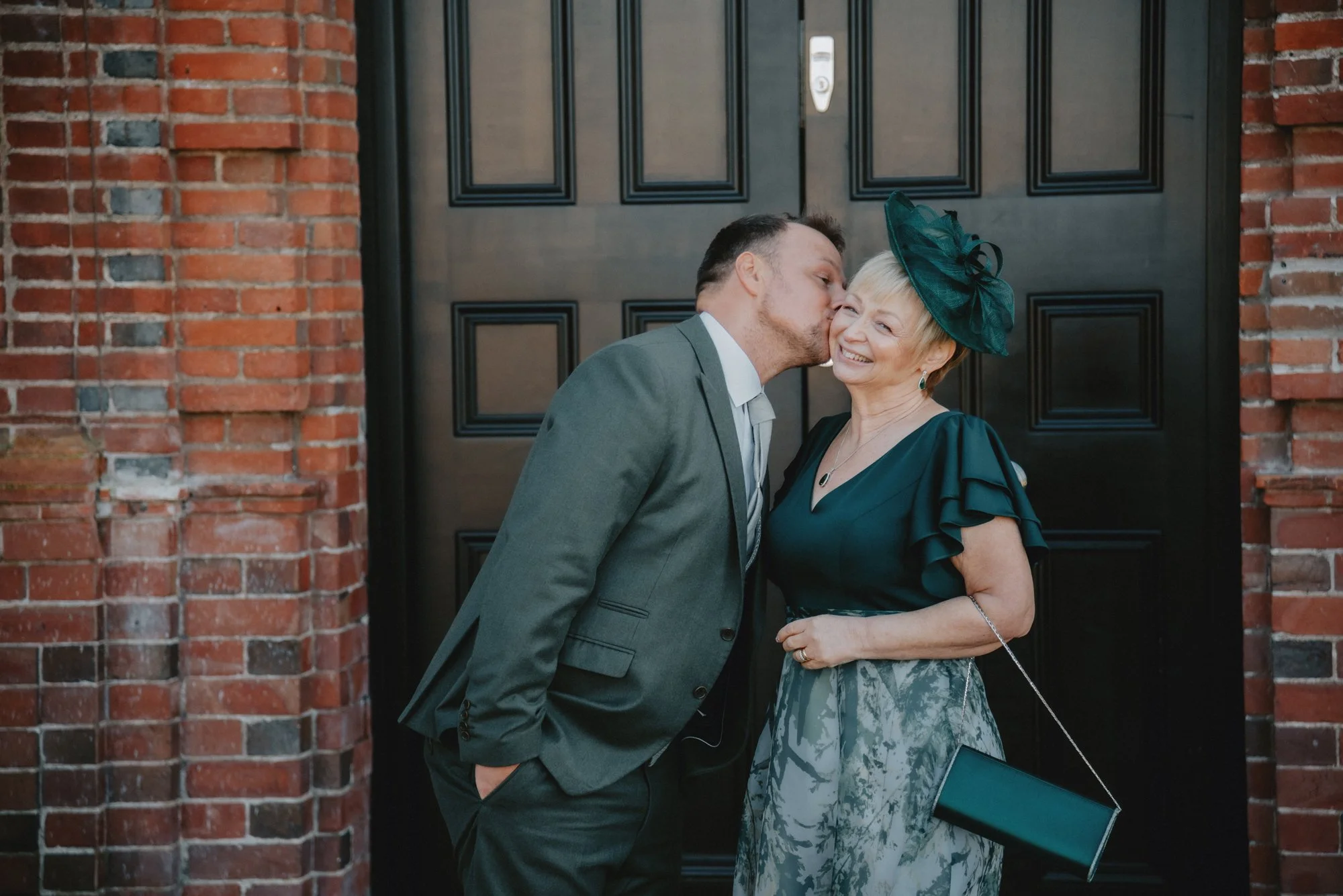 A man in a gray suit kisses an older woman in a teal dress and matching hat on the cheek, standing in front of a black door with brick walls on either side.