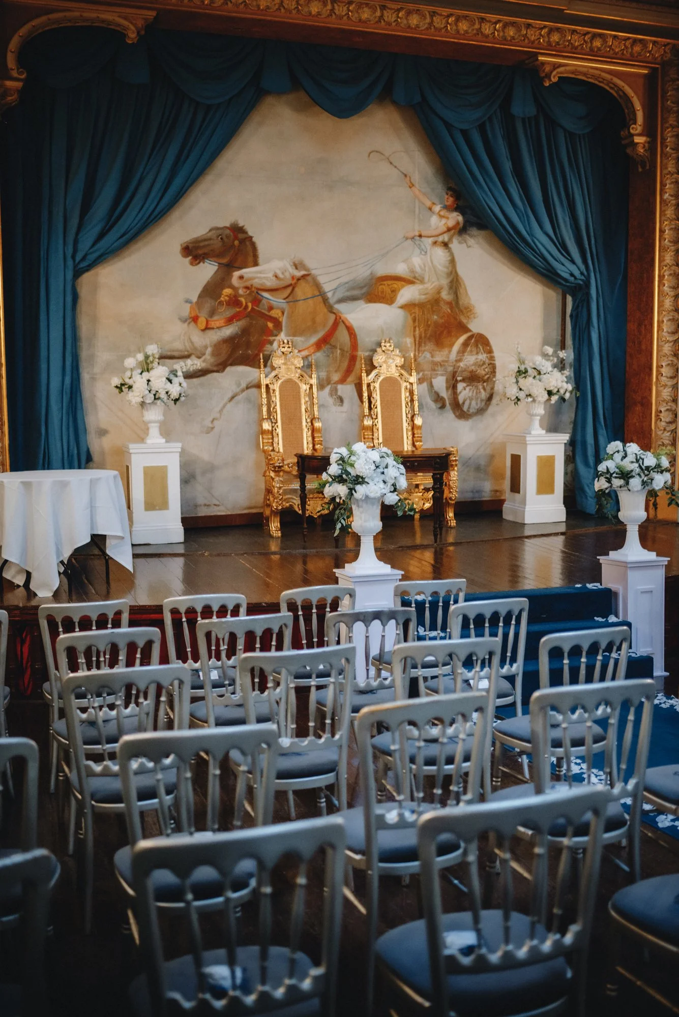 An ornate stage set with a large mural of a chariot drawn by horses, a woman riding it, and blue curtains framing the scene, with white floral arrangements and gold chairs in front for an event.