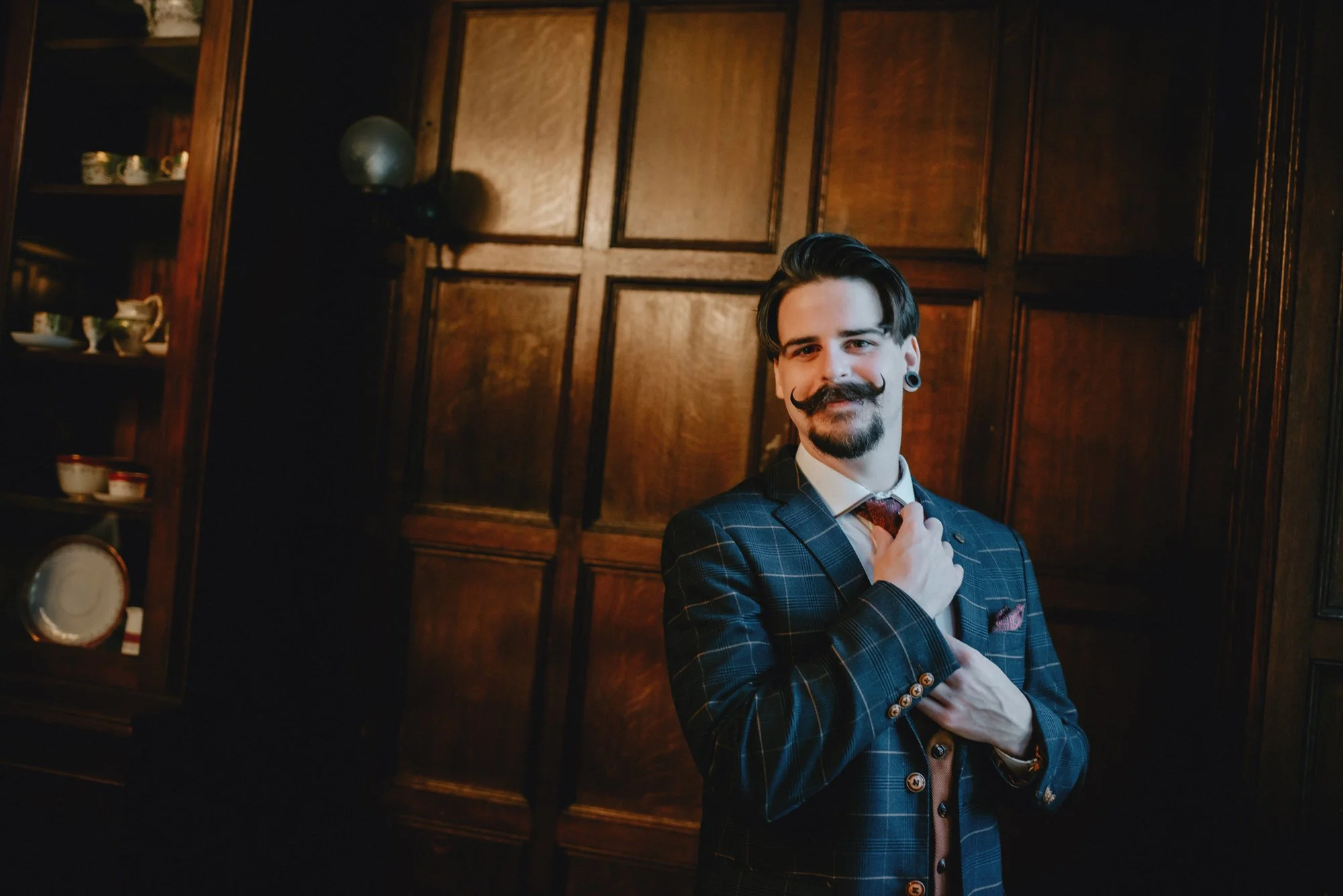 A man with a styled mustache, dark hair, and gauged ears wears a checkered suit, adjusting a tie, standing in front of wooden-paneled wall in a room with china on shelves.