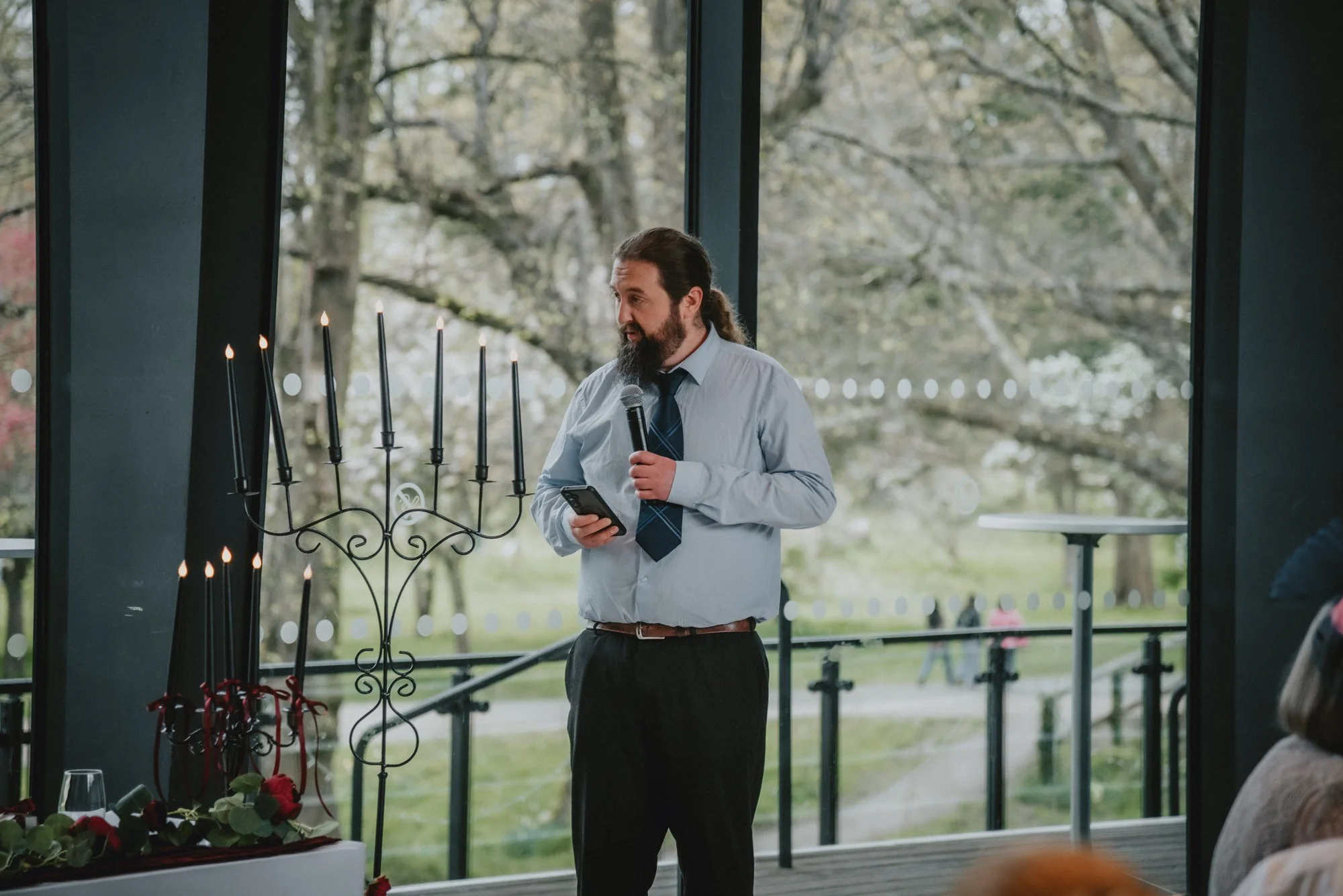 A man with long hair and a beard, wearing a light blue shirt and dark tie, speaking into a microphone while looking at a smartphone during a ceremony in a venue with large glass windows revealing trees outside.