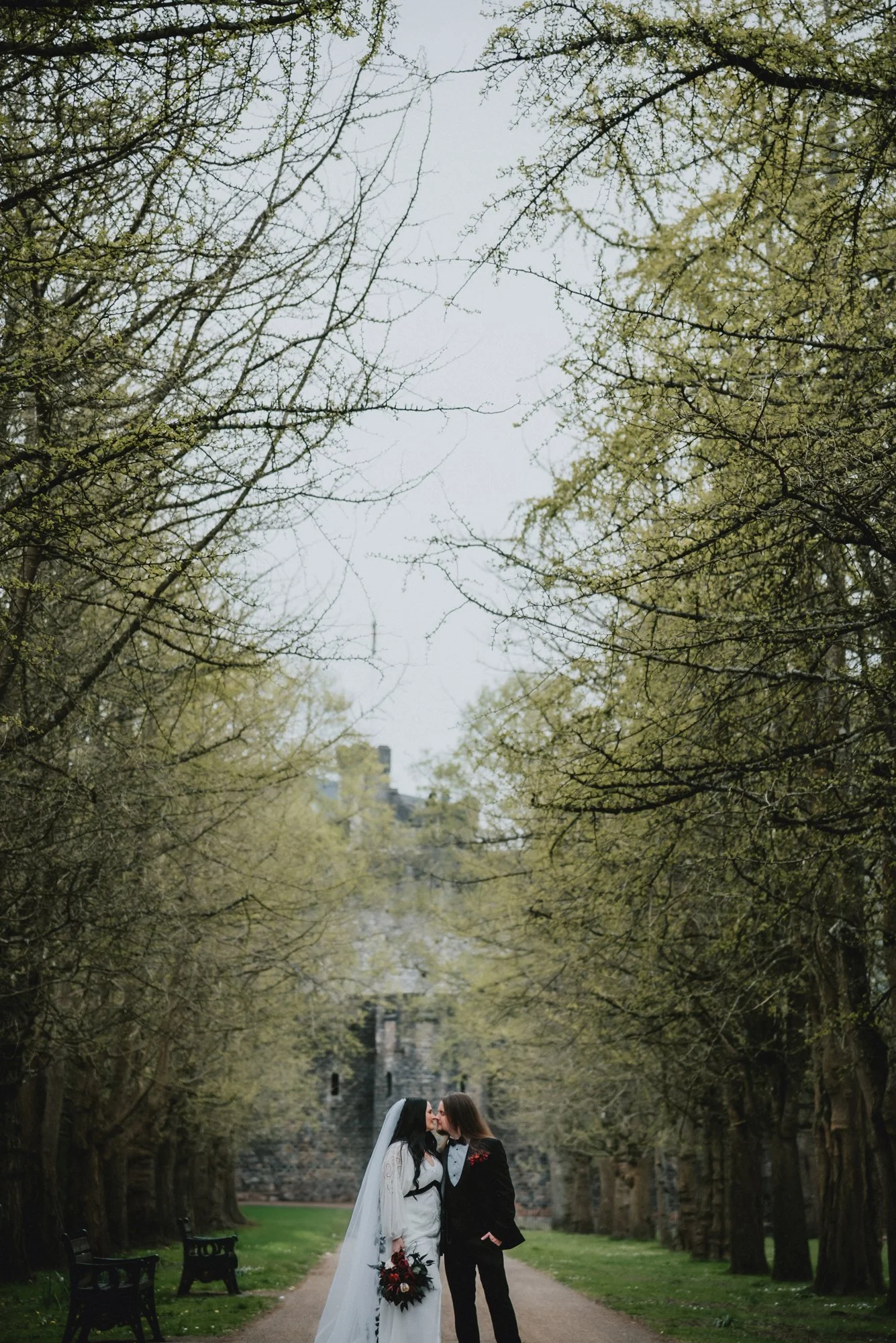 A bride and groom share a kiss on a tree-lined path in a park, with trees on either side and benches along the walkway.