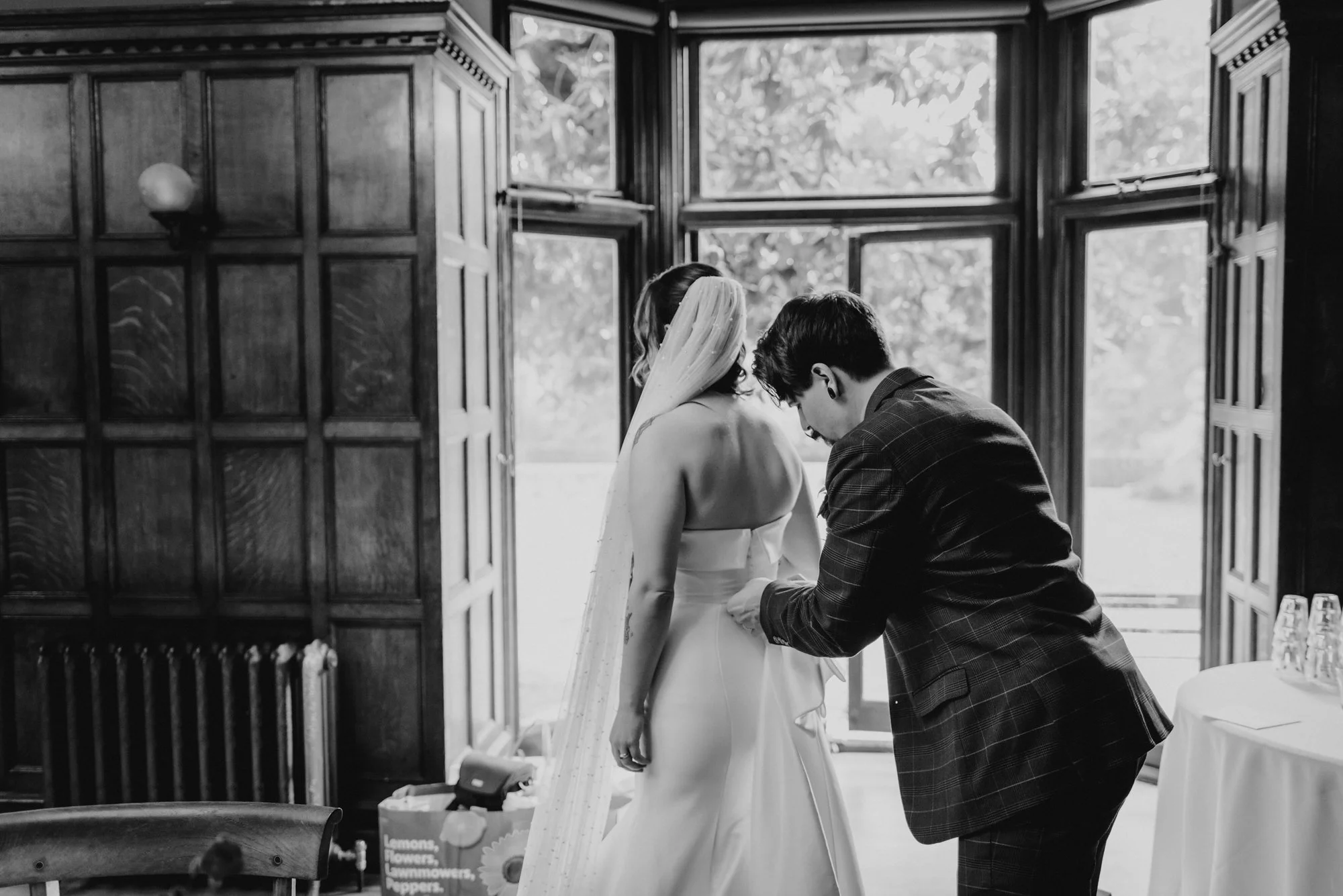A bride in a wedding gown with a veil is being assisted by a man in a suit, possibly during a wedding preparation, standing near large windows with wooden paneling.