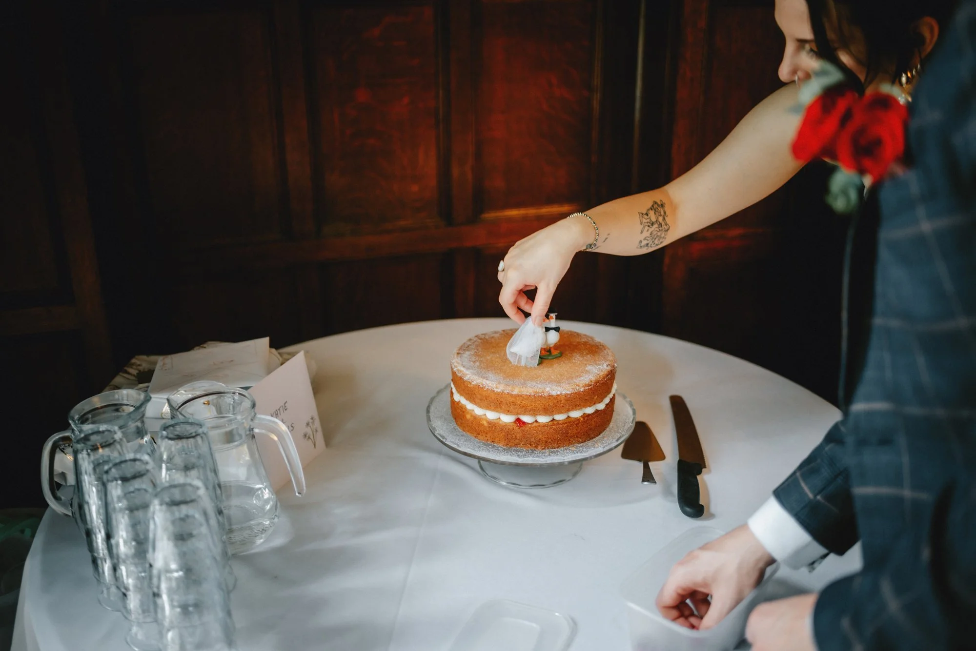 A woman in a gift dress garnished with a rose and leaf in her right hand, is decorating a vanilla layered cake with white frosting on a cake stand, on a white tablecloth.