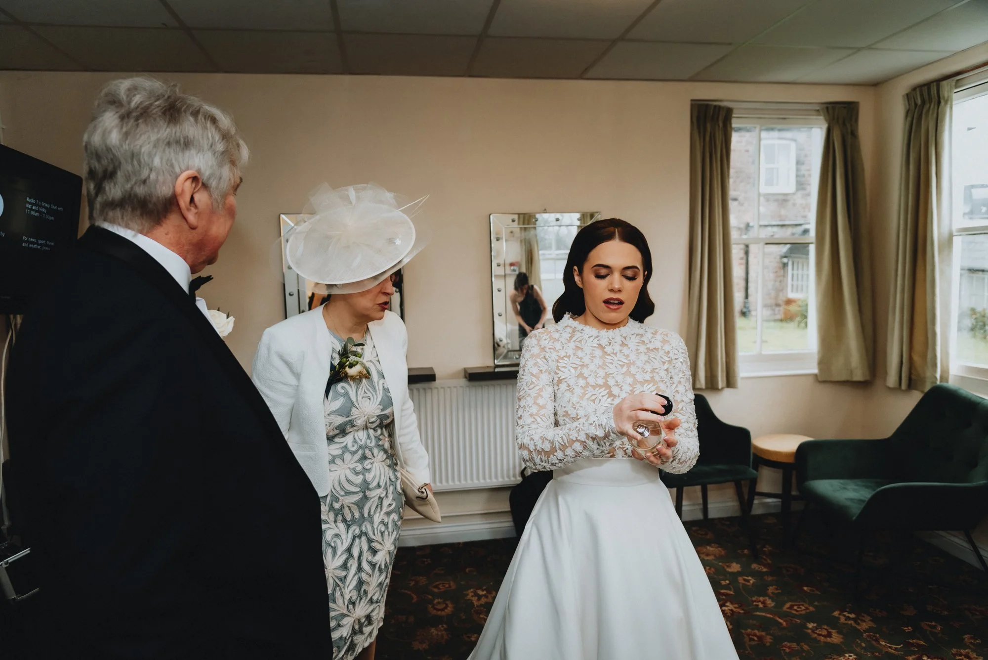 A woman in a white lace top and white skirt preparing for a wedding, with two older adults, a man in a black tuxedo and a woman in a floral dress and white jacket, in a room with beige walls, mirrors, and green chairs.