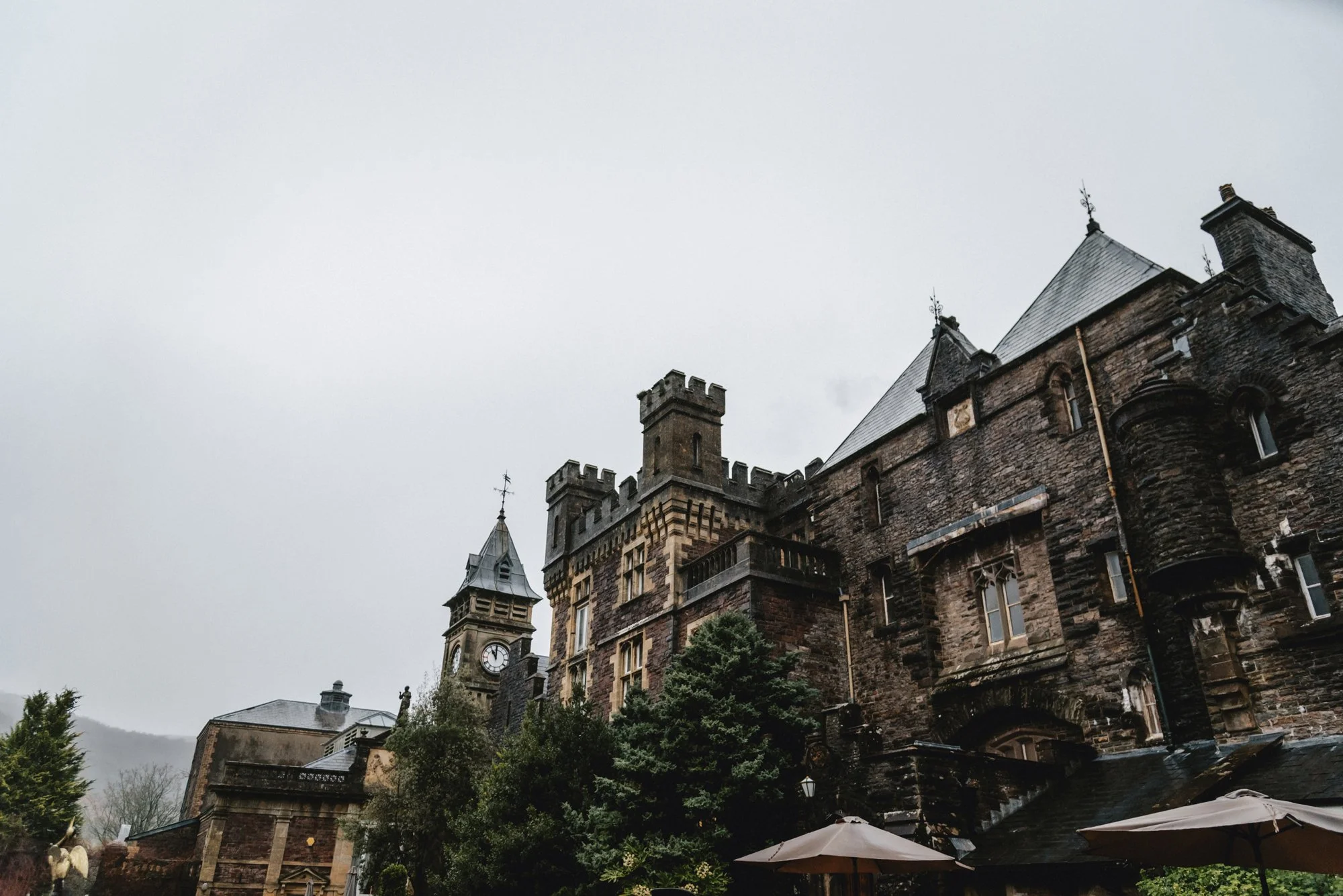 A large, historic castle or mansion made of dark stone with multiple towers and turrets, some topped with weather vanes, set against an overcast sky. There are trees and outdoor umbrellas in the foreground.