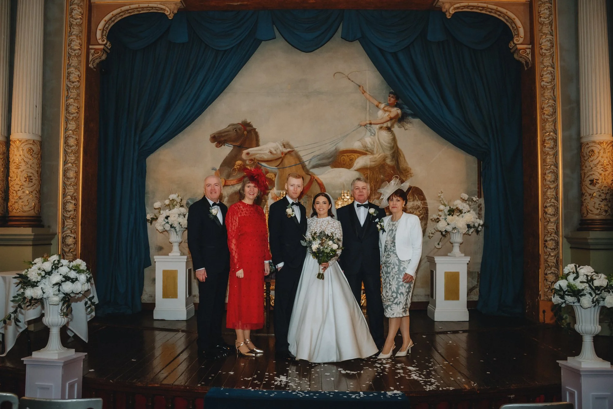 A wedding party standing on a stage in front of a large painting of a chariot with a woman riding it, surrounded by blue curtains, white flowers, and gold ornate decorations.
