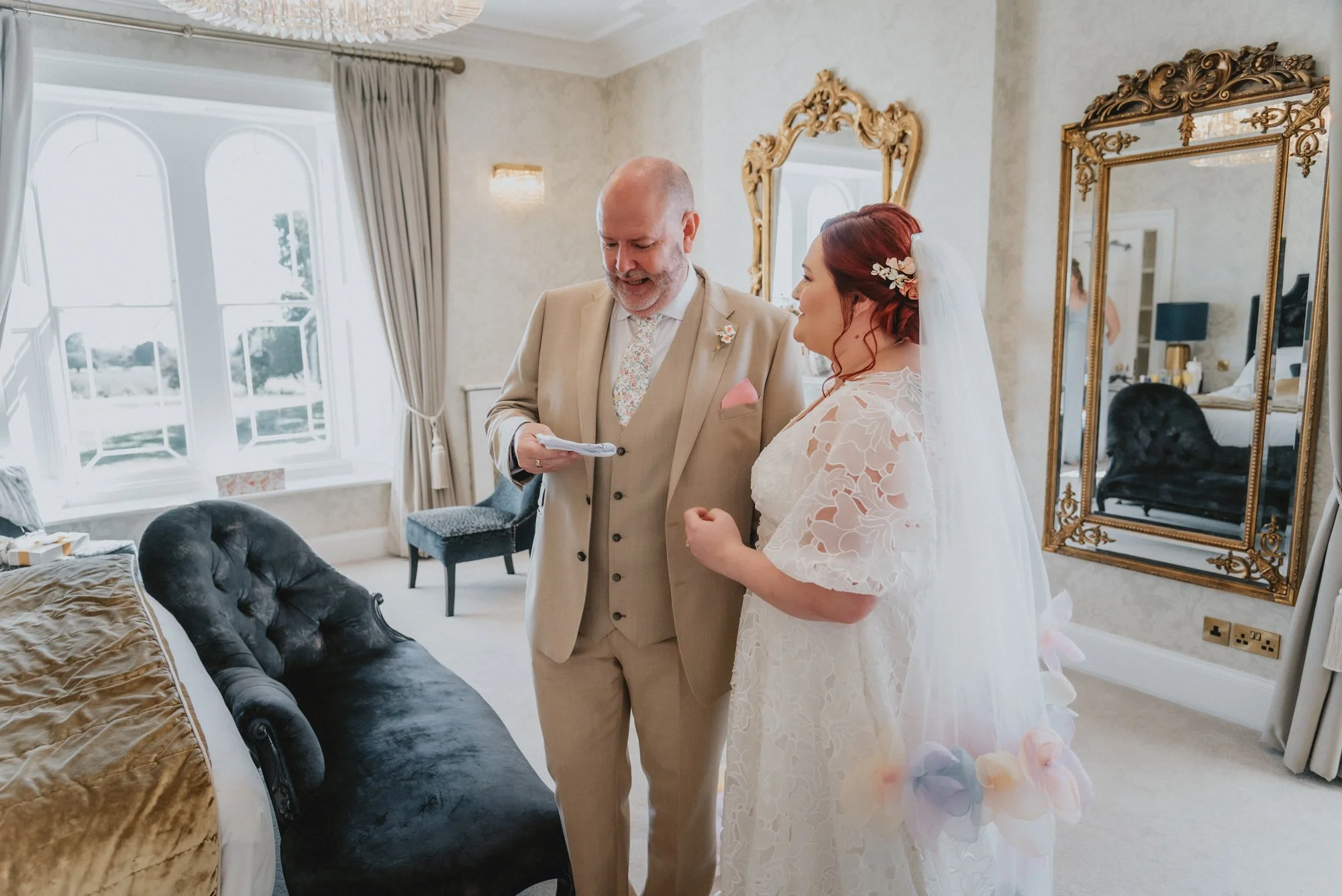 A bride and an older man, likely giving her a speech or reading vows, in a well-lit, elegantly decorated room with large windows, gold ornate mirrors, and vintage furniture.