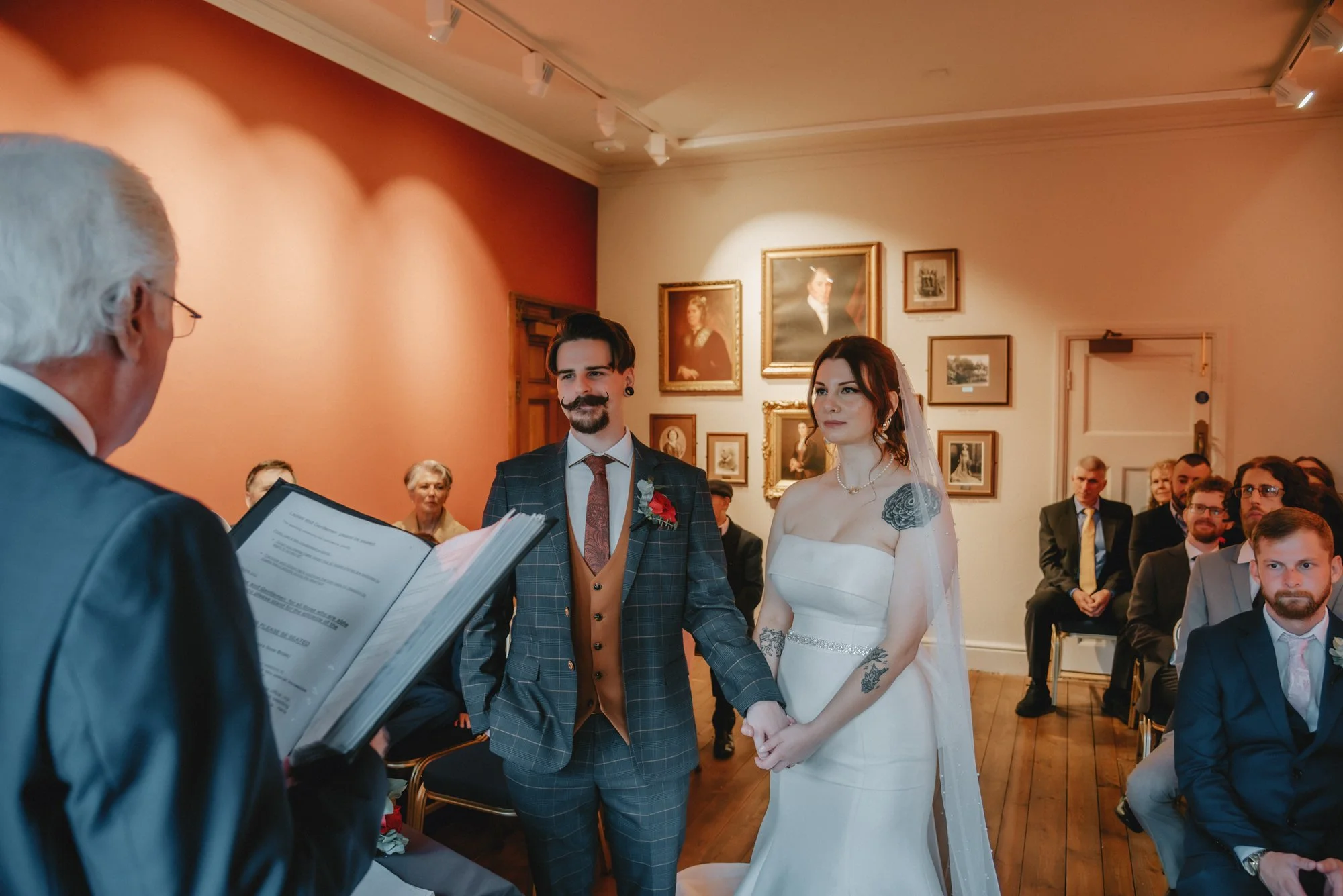A bride and groom standing hand in hand during a wedding ceremony, facing an officiant in a room decorated with framed portraits on the wall, with guests seated in the background.