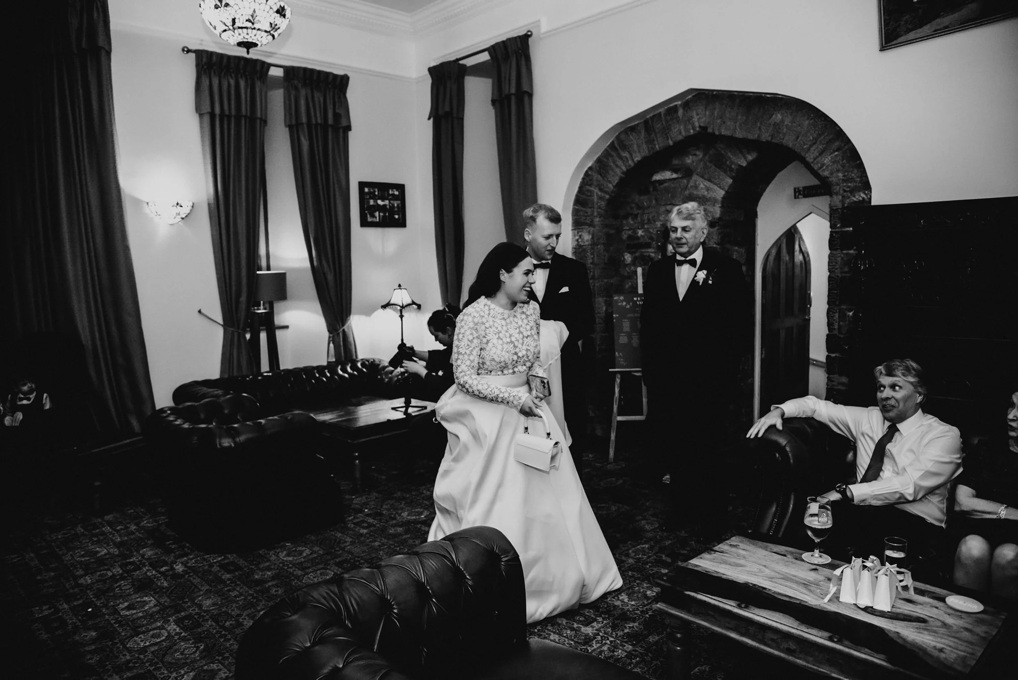 A black and white photo of a wedding reception in a cozy room with dark curtains and vintage furniture. A bride in a long lace dress is smiling, holding a white purse, and standing next to two men in tuxedos. One man is sitting and holding a drink wh
