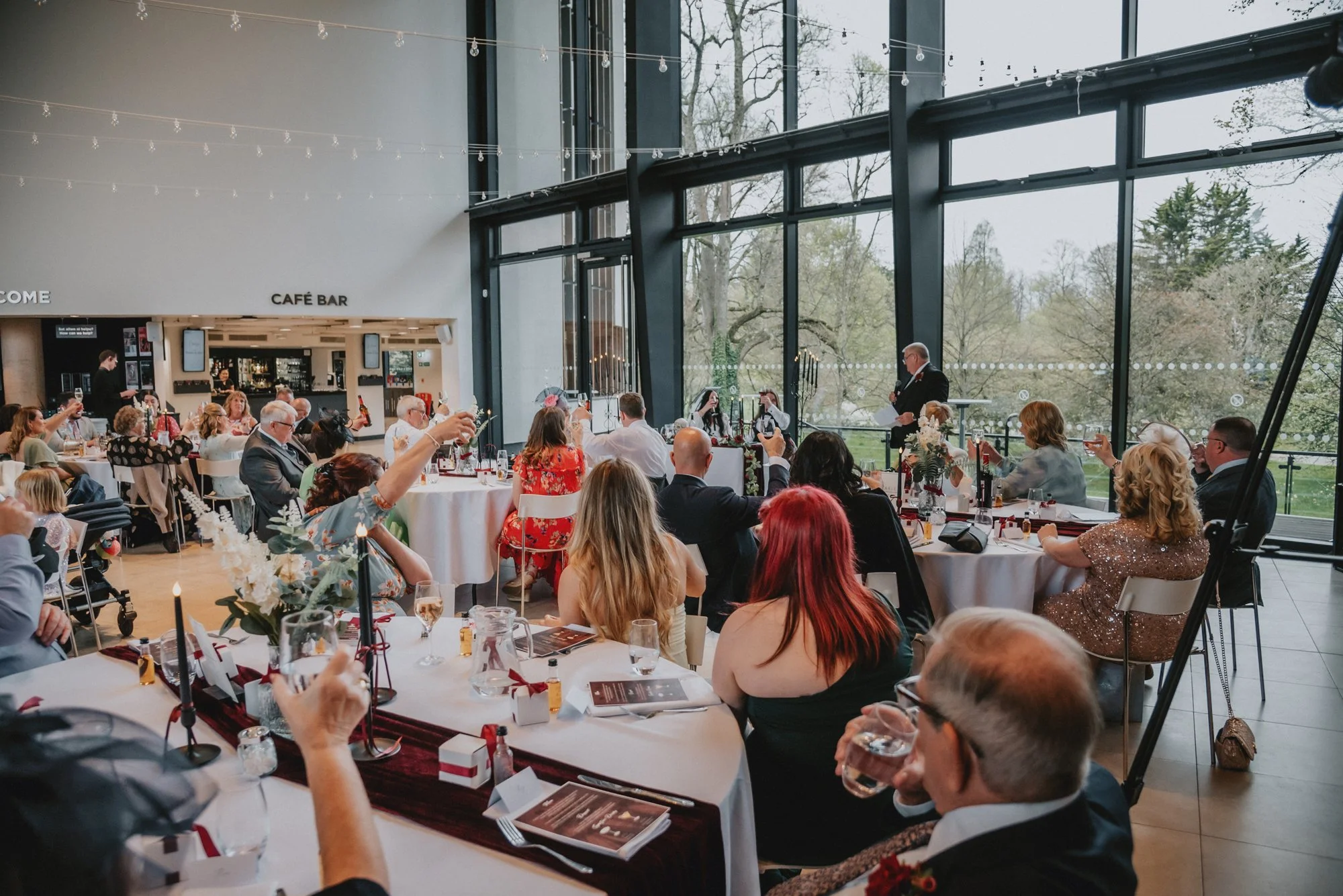 A formal indoor event or celebration with people seated at round tables, some raising glasses, in a bright room with large glass windows overlooking trees. A man in a suit stands at a podium.