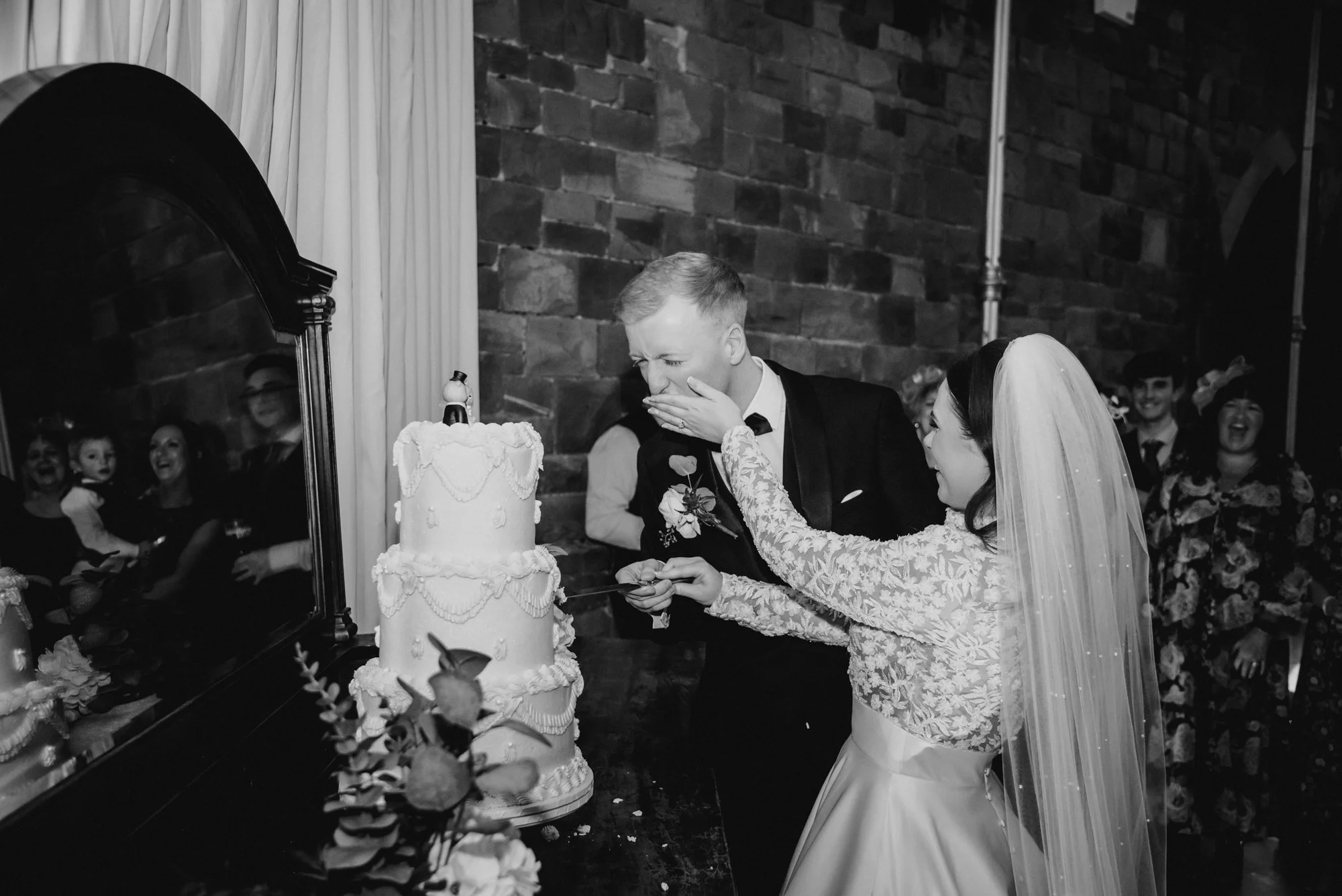 A bride and groom cut their wedding cake together, with the bride playfully covering the groom's mouth. The wedding cake is decorated with icing and flowers, and a mirror reflects the guests watching and smiling.