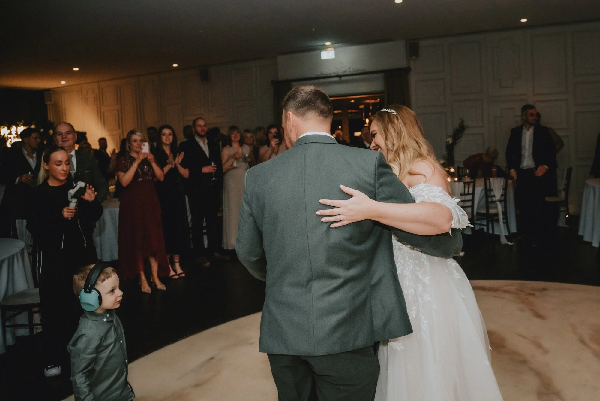 A bride and groom dance together at their wedding reception, surrounded by guests who are clapping and watching in a decorated indoor venue.