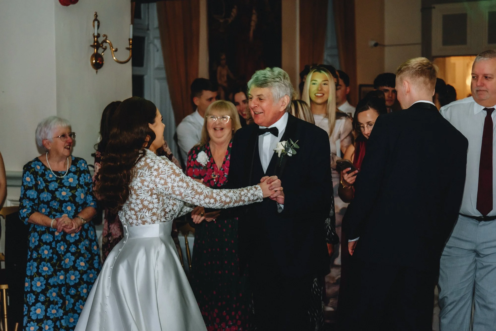 A bride and an older man, possibly her father, dancing and holding hands at a wedding reception, surrounded by smiling guests.