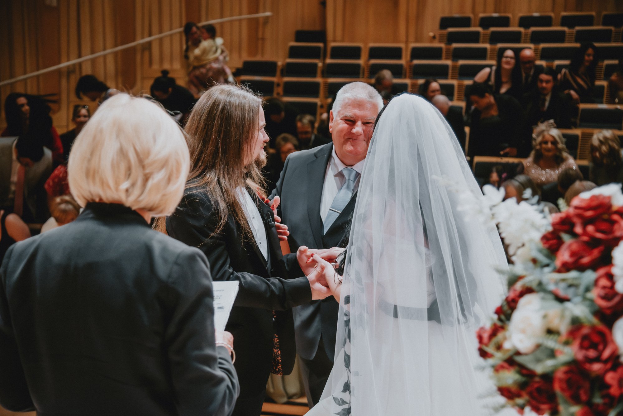 A bride and groom holding hands during their wedding ceremony, facing an officiant, in a parish with wooden walls and seating filled with guests.