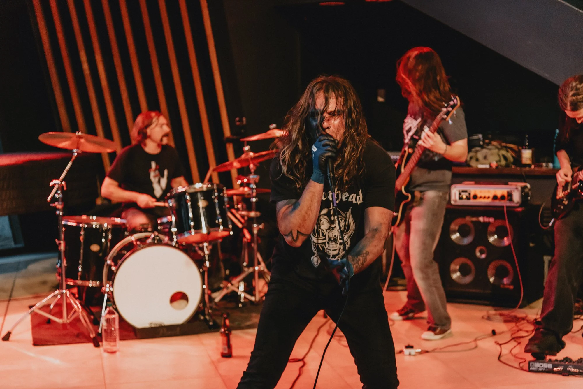 A band performs live on stage with a lead vocalist with long hair, tattoos, and a black T-shirt, along with a drummer and two guitarists, all playing their instruments.
