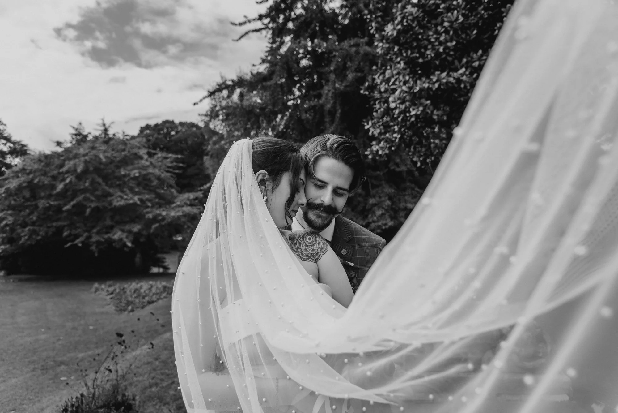 Black and white photo of a bride and groom close together outdoors, with the bride wearing a veil and the groom dressed in a suit, both smiling gently.