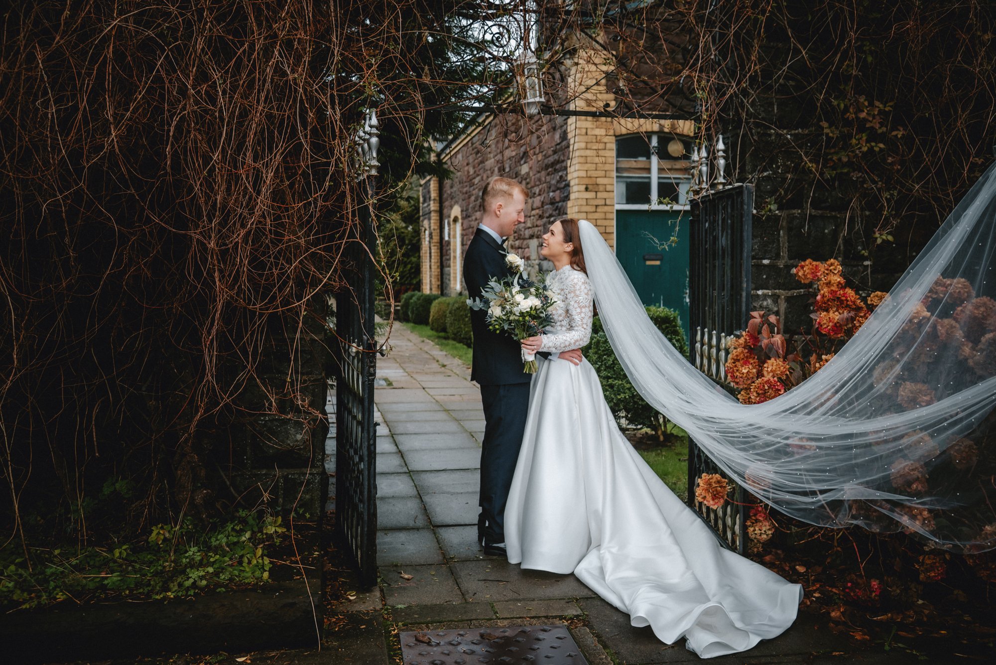 A bride and groom standing close together in a garden entrance, smiling and holding hands. The bride is holding a bouquet and wearing a white wedding gown with a long veil, while the groom is in a black tuxedo. The garden gate is open, showing a bric
