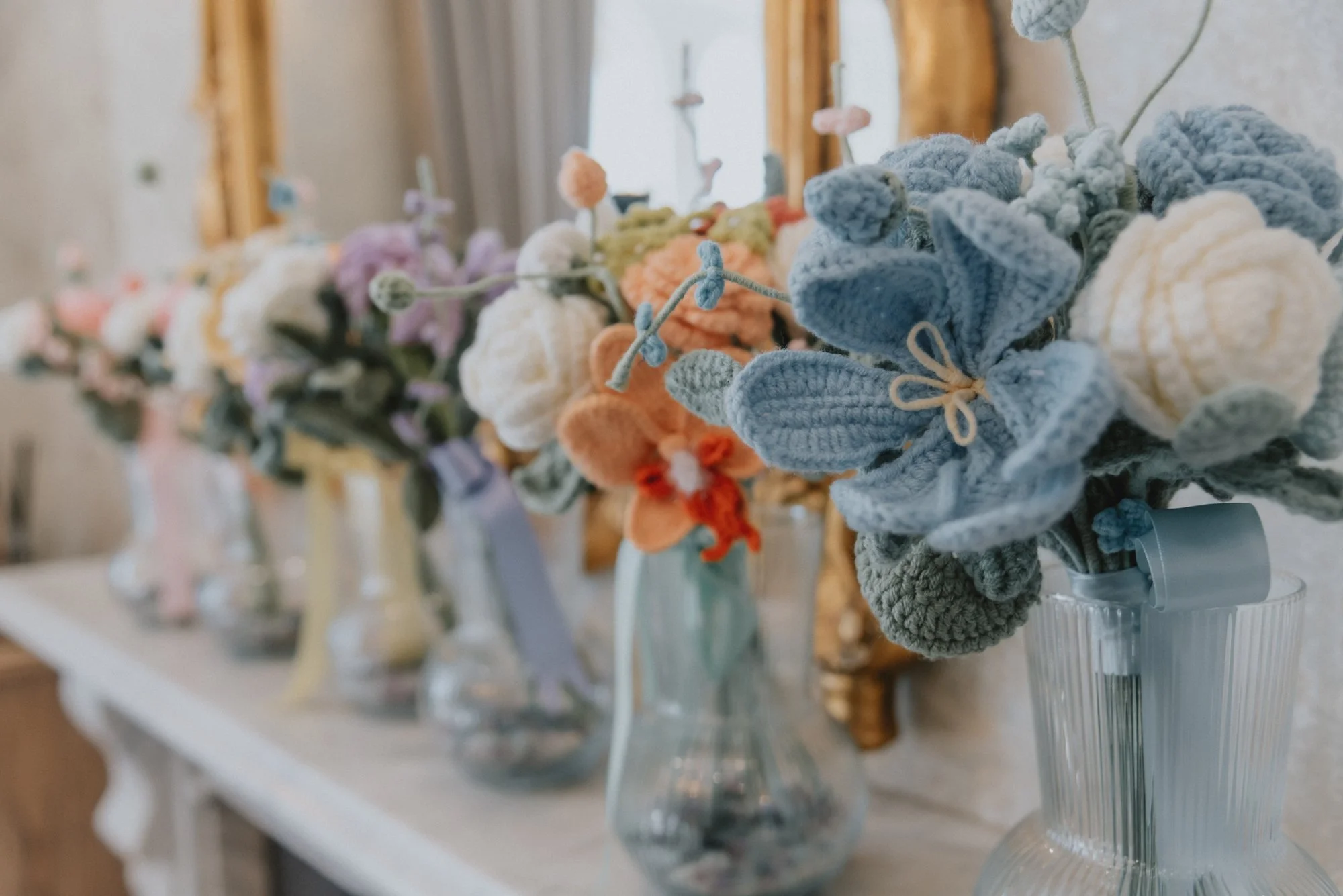 A row of colorful, knitted flower bouquets in glass vases on a white wooden table, with a mirror and curtains in the background.