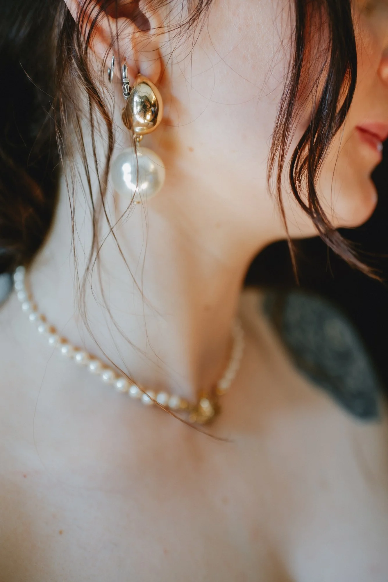 Close-up of a woman's face showing her ear with gold and pearl earrings, part of her face, shoulder, and a tattoo on her shoulder. She is wearing a pearl necklace.