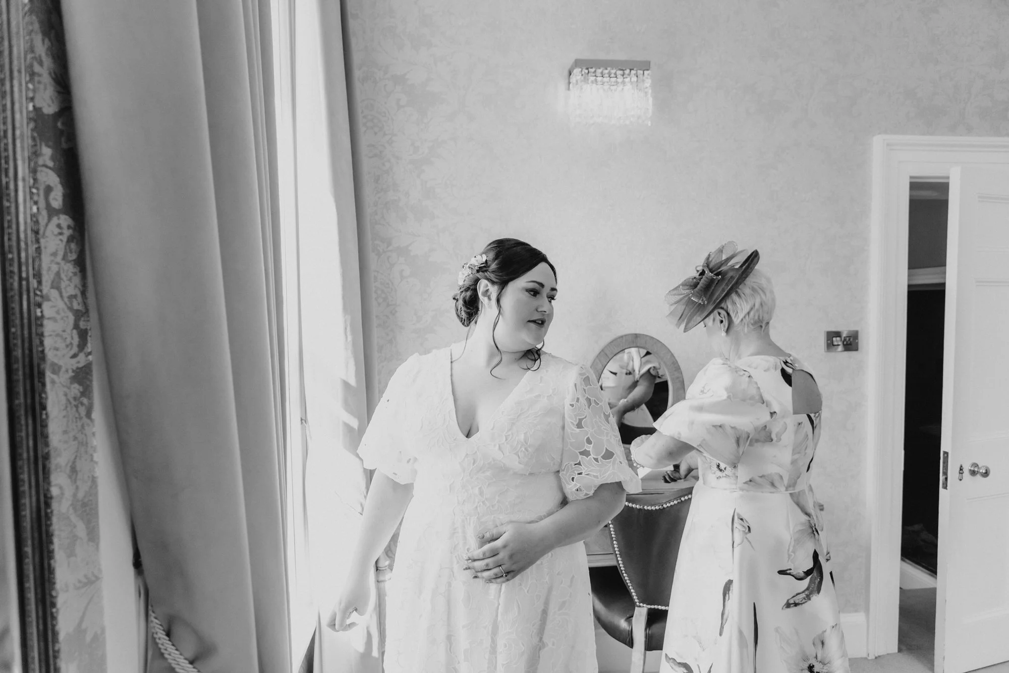 A woman in a lace wedding dress with dark hair styled in an updo, standing in a room, talking to an older woman with a short blonde hairstyle wearing a patterned dress and a fascinator hat. The room has curtains, a mirror on a dresser, and wallpaper.