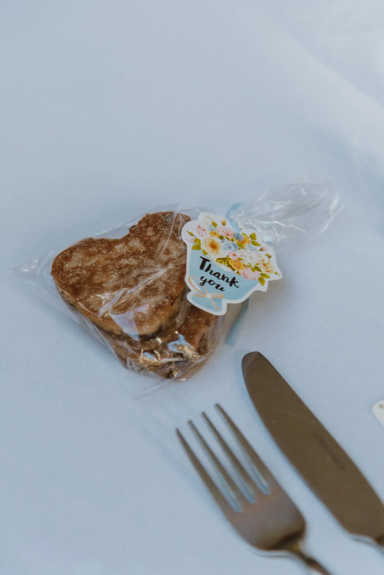 Wrapped heart-shaped baked good with a 'Thank you' sticker on a white tablecloth, next to a fork and knife.