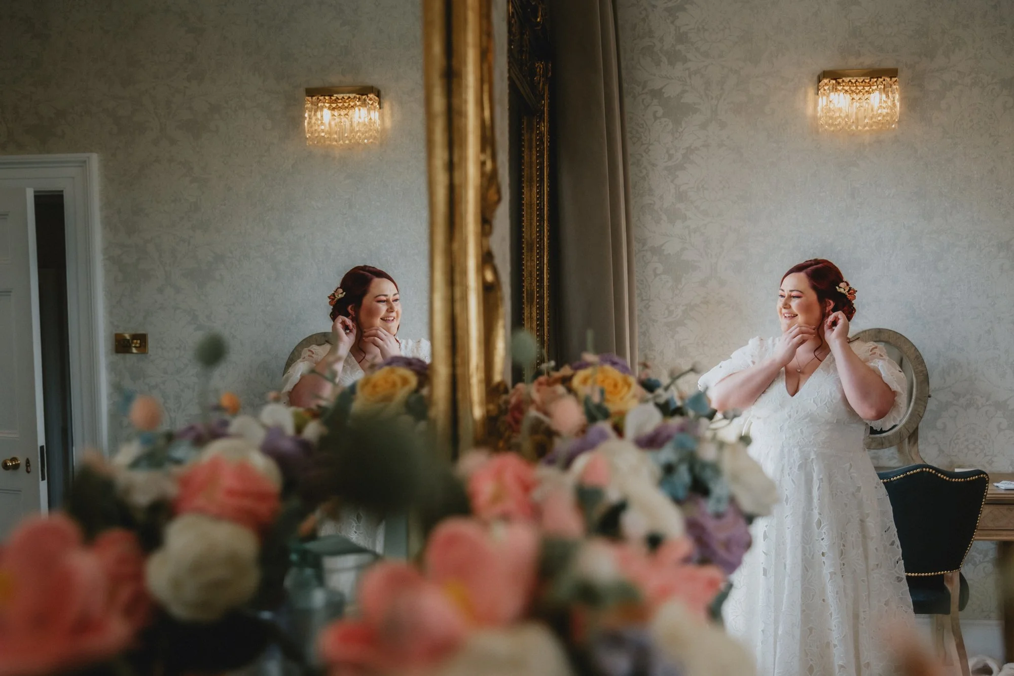A bride in a lace wedding dress smiling and adjusting her earring in front of a mirror, with a floral arrangement in the foreground and elegant chandelier lighting in the background.