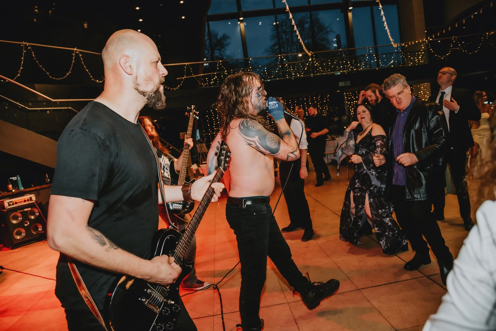 Band performing at a dance party with a guitarist, singer with tattoos, and audience dancing under string lights in a large indoor venue.