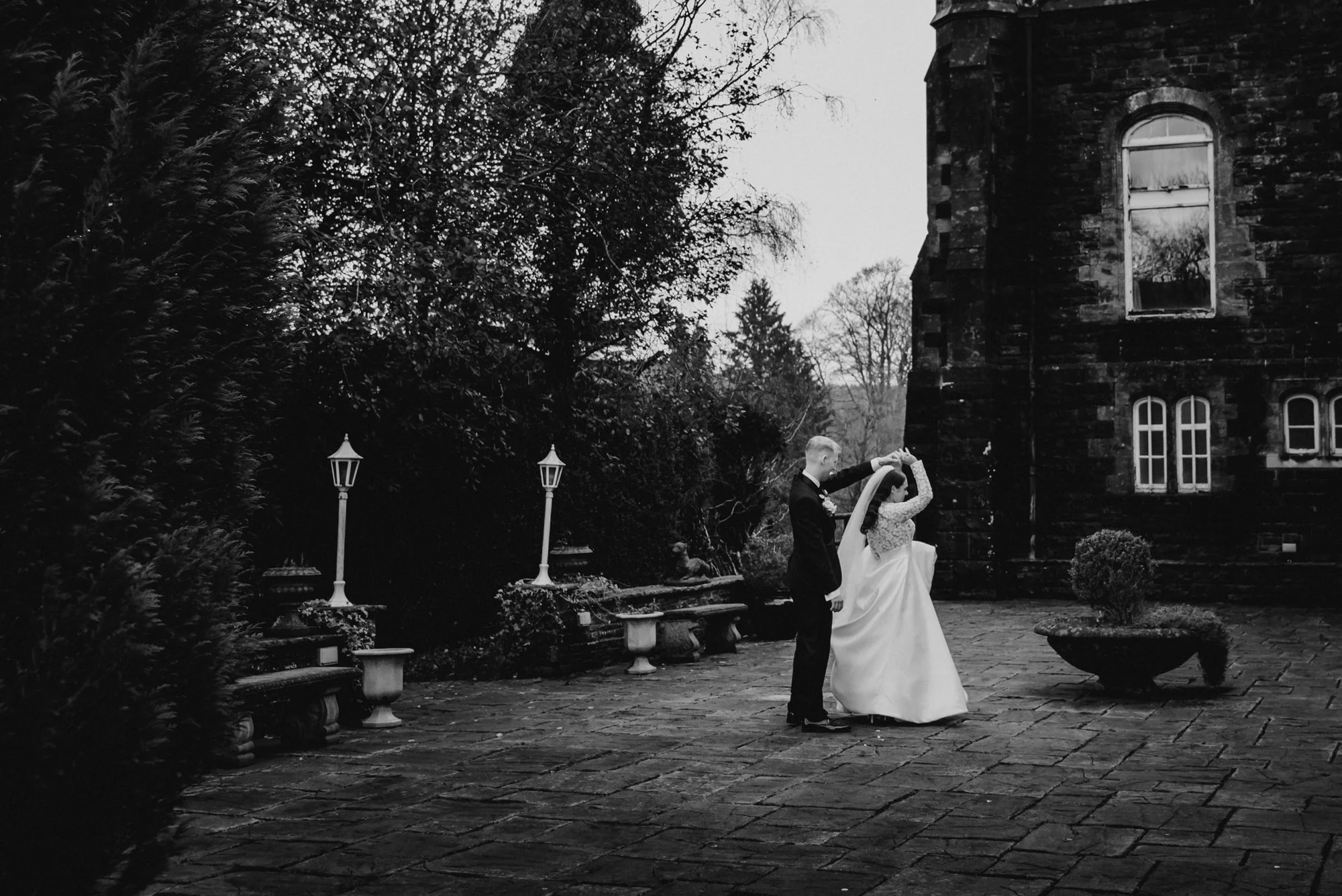 A black and white photo of a bride and groom dancing outdoors near an old brick building with windows. The groom in a suit holds the bride's hand as she joins him in dance on a stone patio, with trees, benches, and decorative street lamps in the back