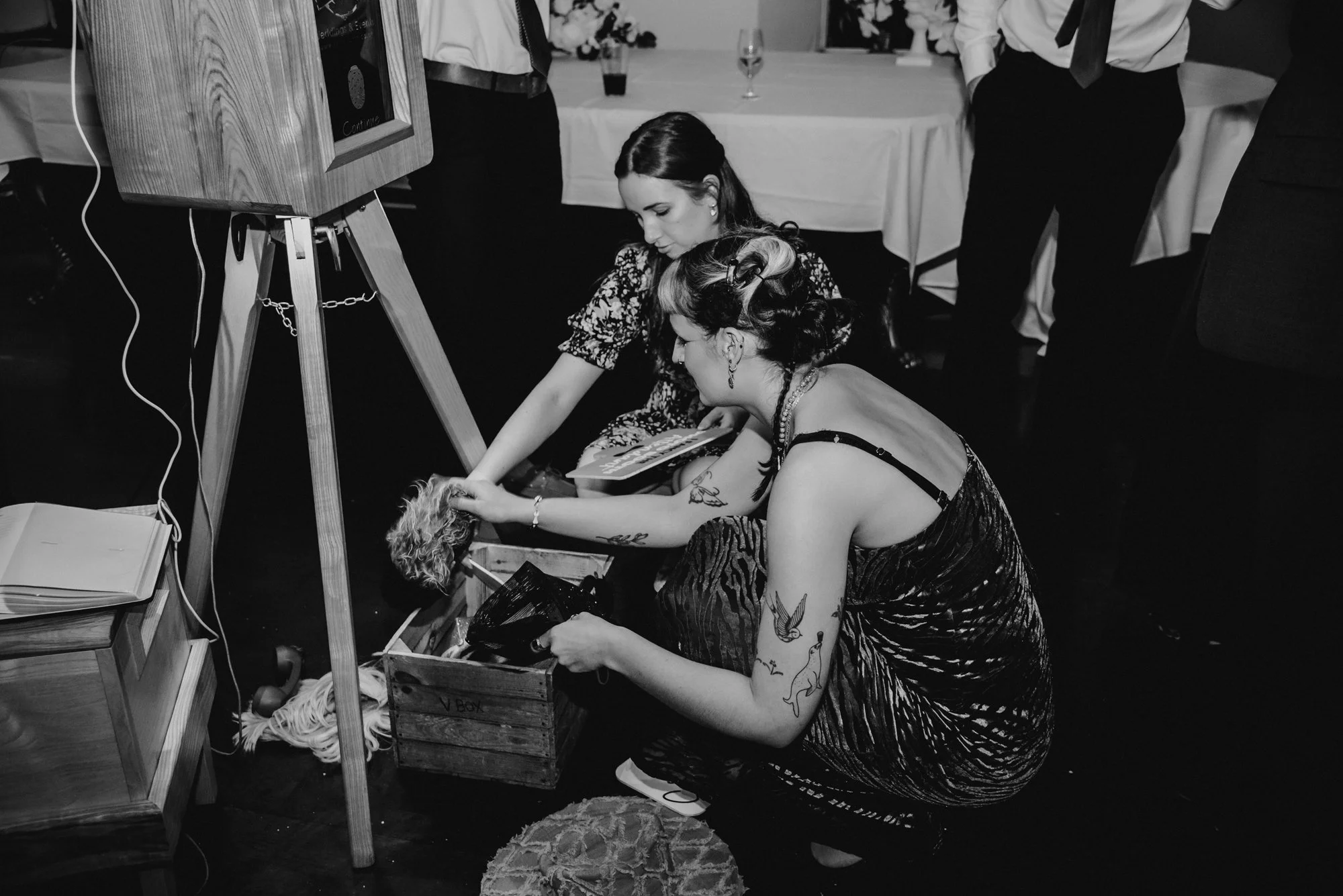 Two women with tattoos and styled hair are kneeling on the floor, looking through a wooden box with various items inside at a formal event with tables, white tablecloths, and cloudy drinks in the background.