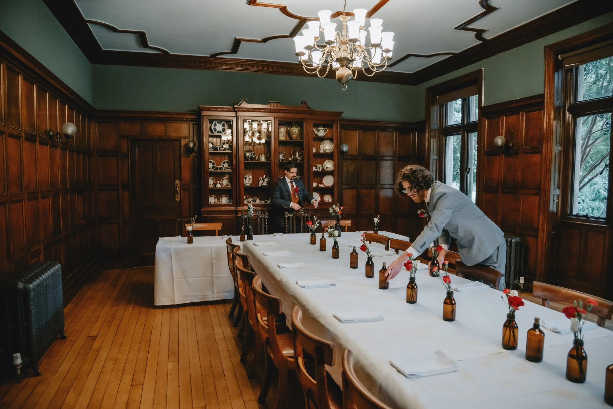 A man in a gray suit arranging flowers in small brown bottles on a dining table in a wood-paneled room with large windows and a chandelier.