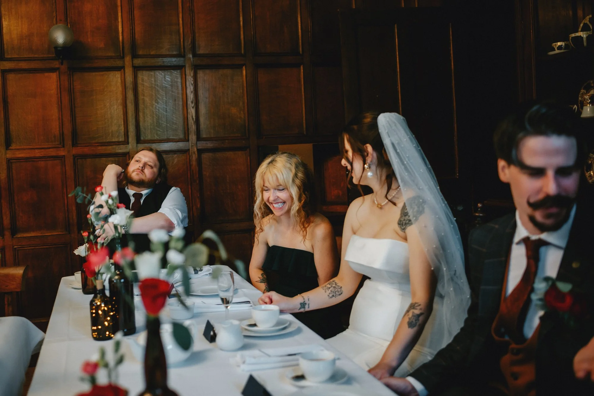 A wedding reception with four people sitting at a table, including a bride in a white dress and veil, two women, and a man, all laughing or smiling. The table is decorated with flowers and set with cups and plates. The background features wooden pane