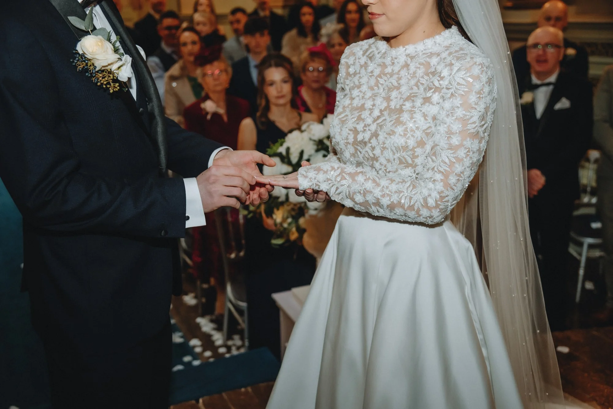 A bride and groom during their wedding ceremony, holding hands, with guests watching in the background.