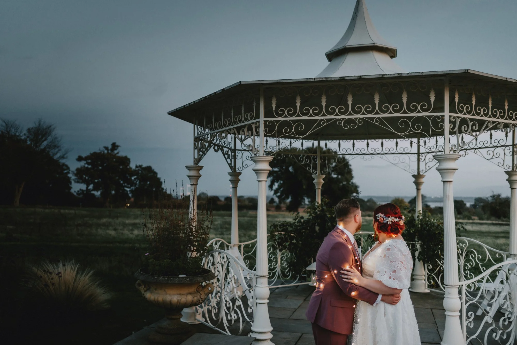 A couple dressed in wedding attire standing close together under a decorative white gazebo outdoors during dusk.