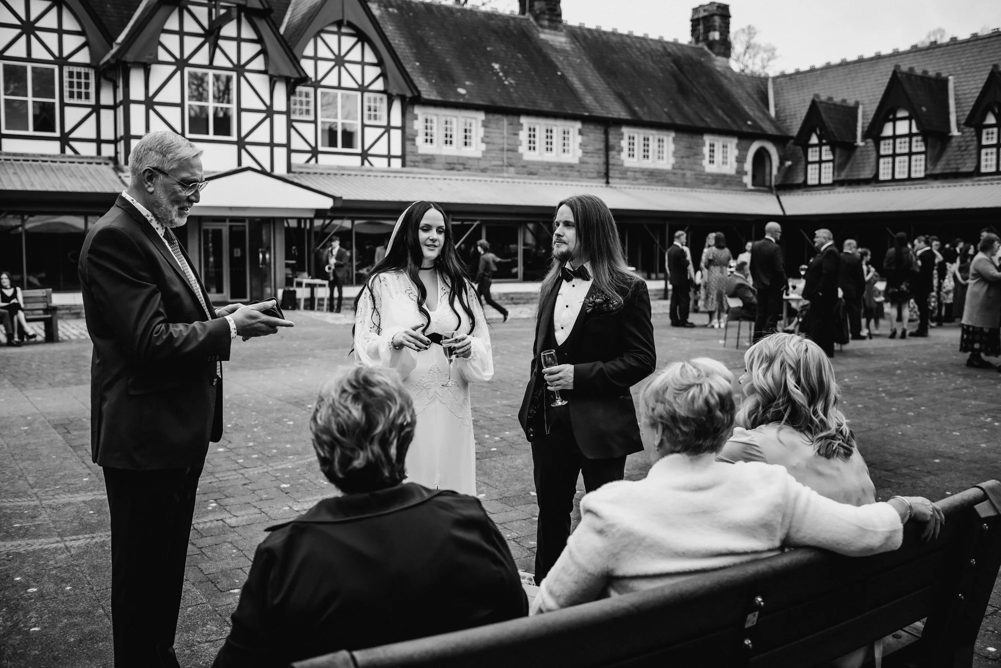 People gathered at an outdoor event, with a man using a mobile phone, a woman and man in formal attire holding drinks, and several others sitting on a bench, with a large building with Tudor-style architecture in the background.