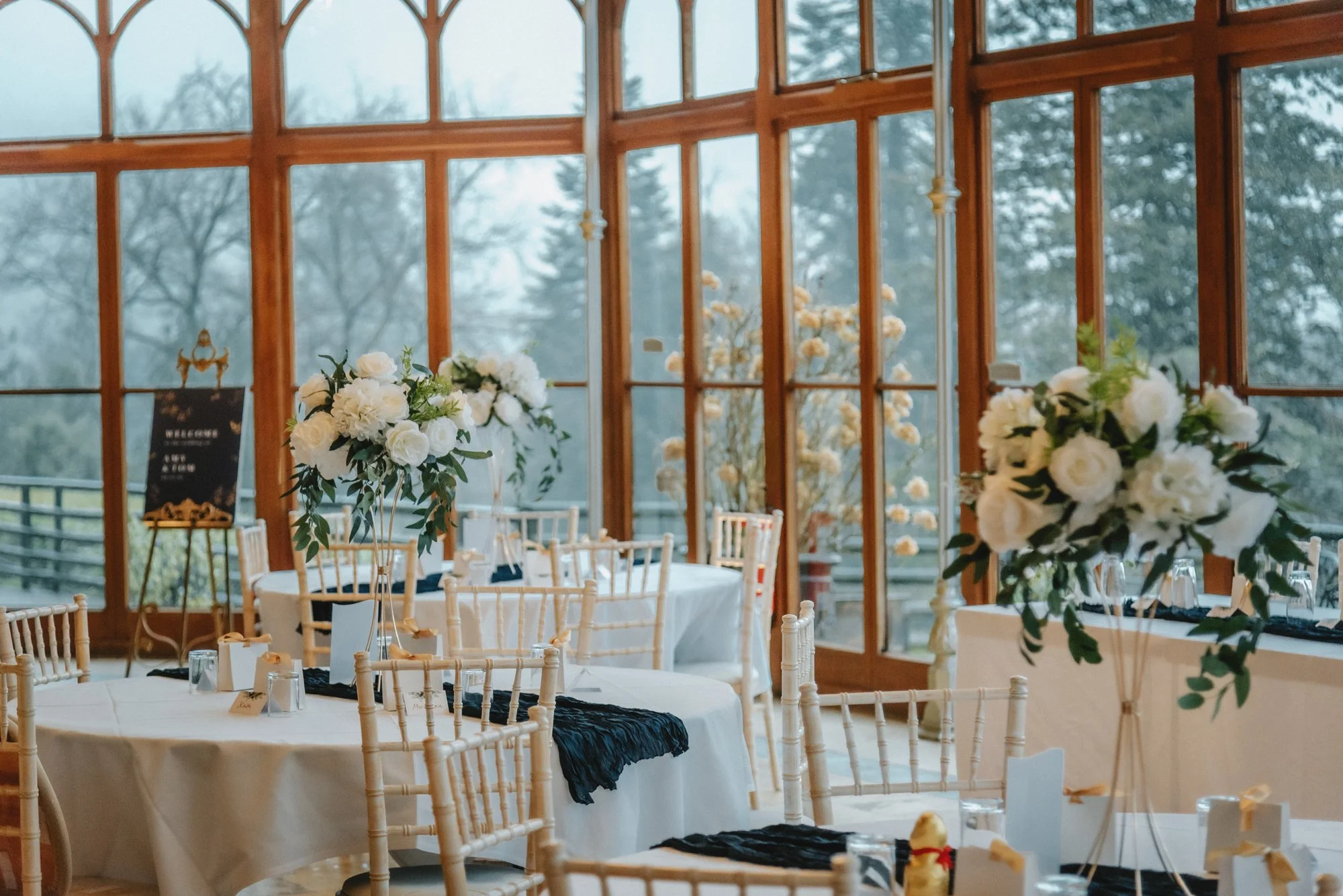 An elegant event space with round tables covered in white tablecloths, decorated with tall floral centerpieces of white roses and greenery, and surrounded by white chairs. The background features large wooden-framed windows with a view of trees outsi