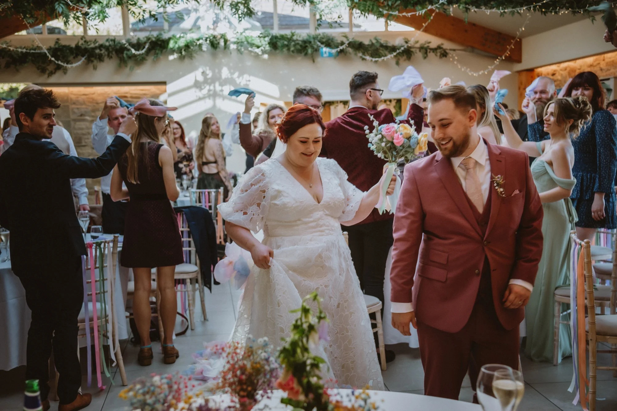 A bride and groom at their wedding reception, engaged in a celebratory dance, surrounded by wedding guests in a decorated venue with string lights and floral decorations.