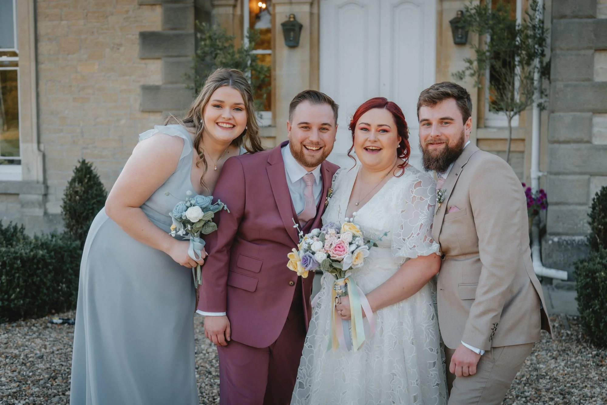 Group of five people smiling at a wedding, two women in dresses holding bouquets, two men in suits, and a bride in a white gown holding a bouquet, standing outside a building with brick walls and plants.