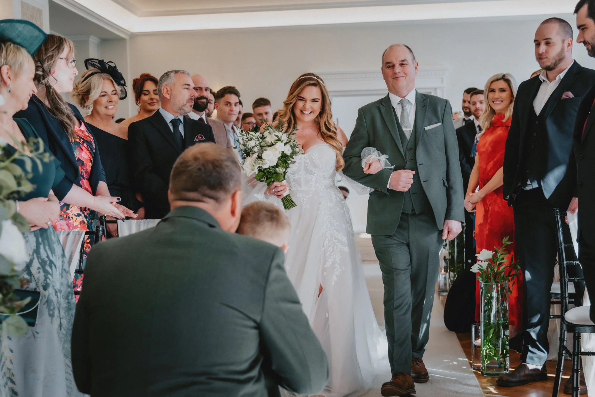 Bride with a bouquet walking down the aisle accompanied by an older man, surrounded by guests at a wedding ceremony in a bright room.
