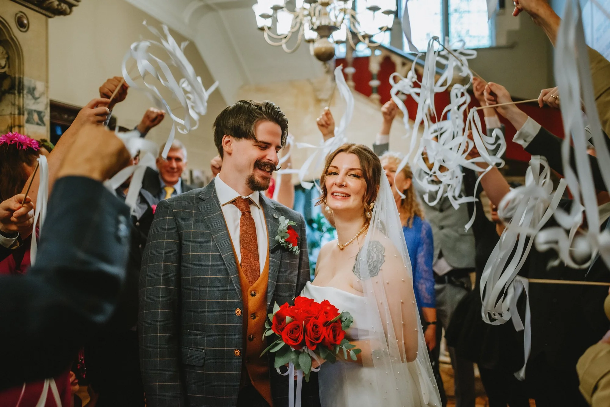 A newlywed couple stands under a shower of white streamers, smiling. The bride is holding a bouquet of red roses and wearing a wedding dress and veil, while the groom is dressed in a checked suit with a brown vest and tie. Guests surround them celebr