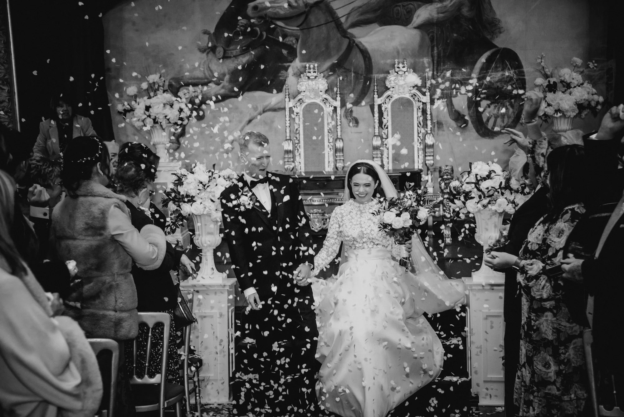 A black and white photograph of a wedding ceremony with a bride and groom walking down the aisle, surrounded by guests, as flower petals are thrown in the air.